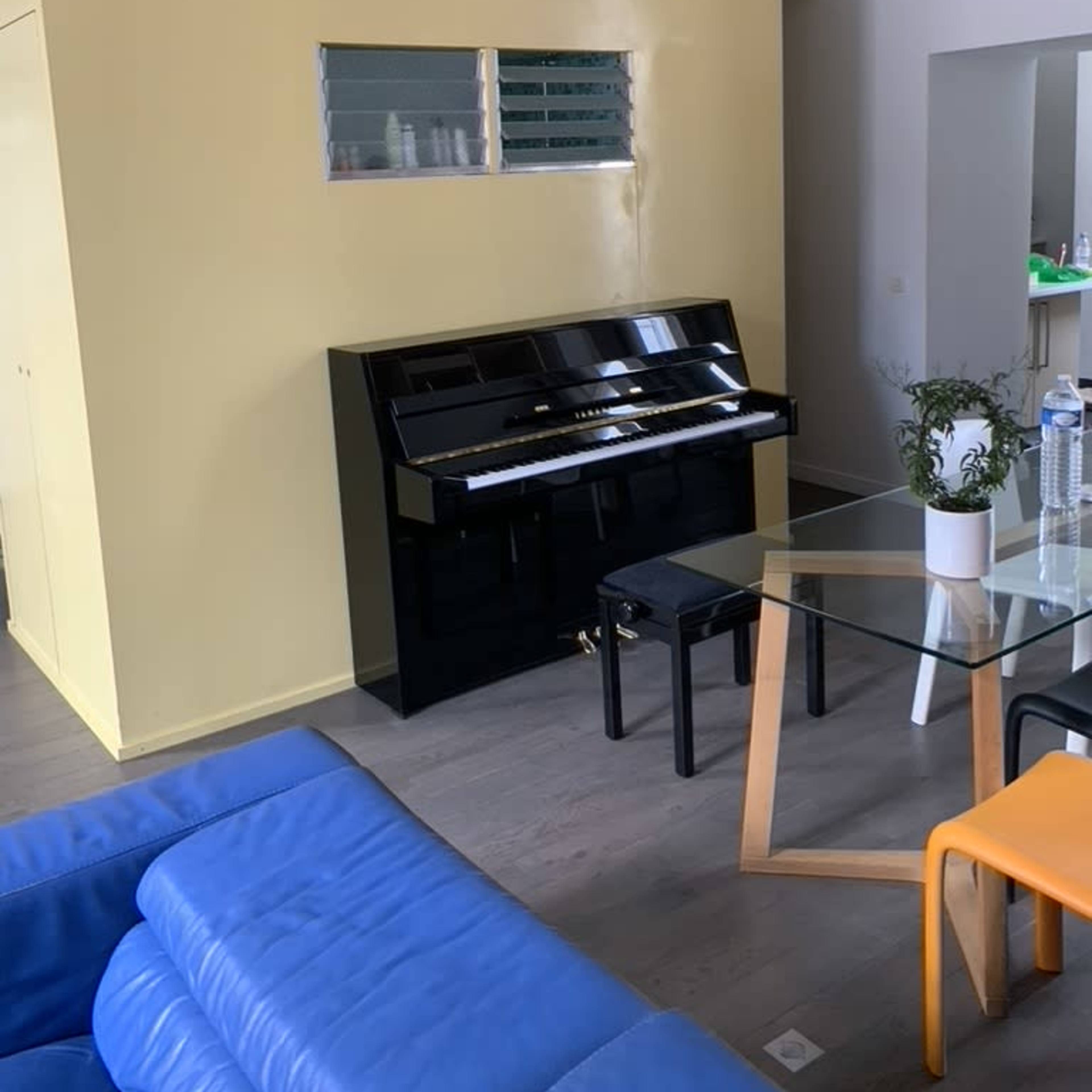 A black piano sits against a yellow wall in a room furnished with a glass table and blue and orange chairs.
