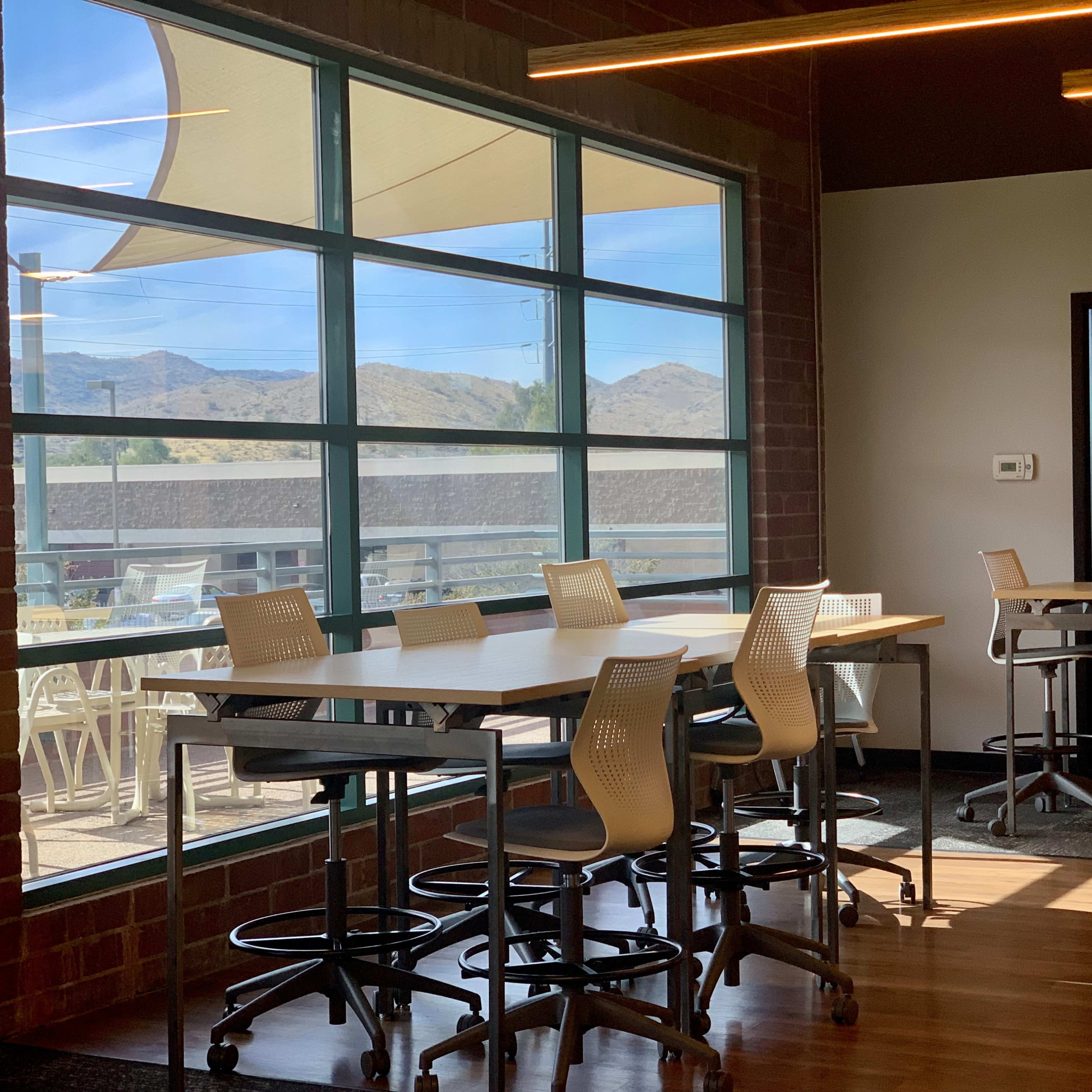 A bright, modern workspace featuring several high tables with stools beside large windows that overlook a scenic outdoor area with mountains in the background.