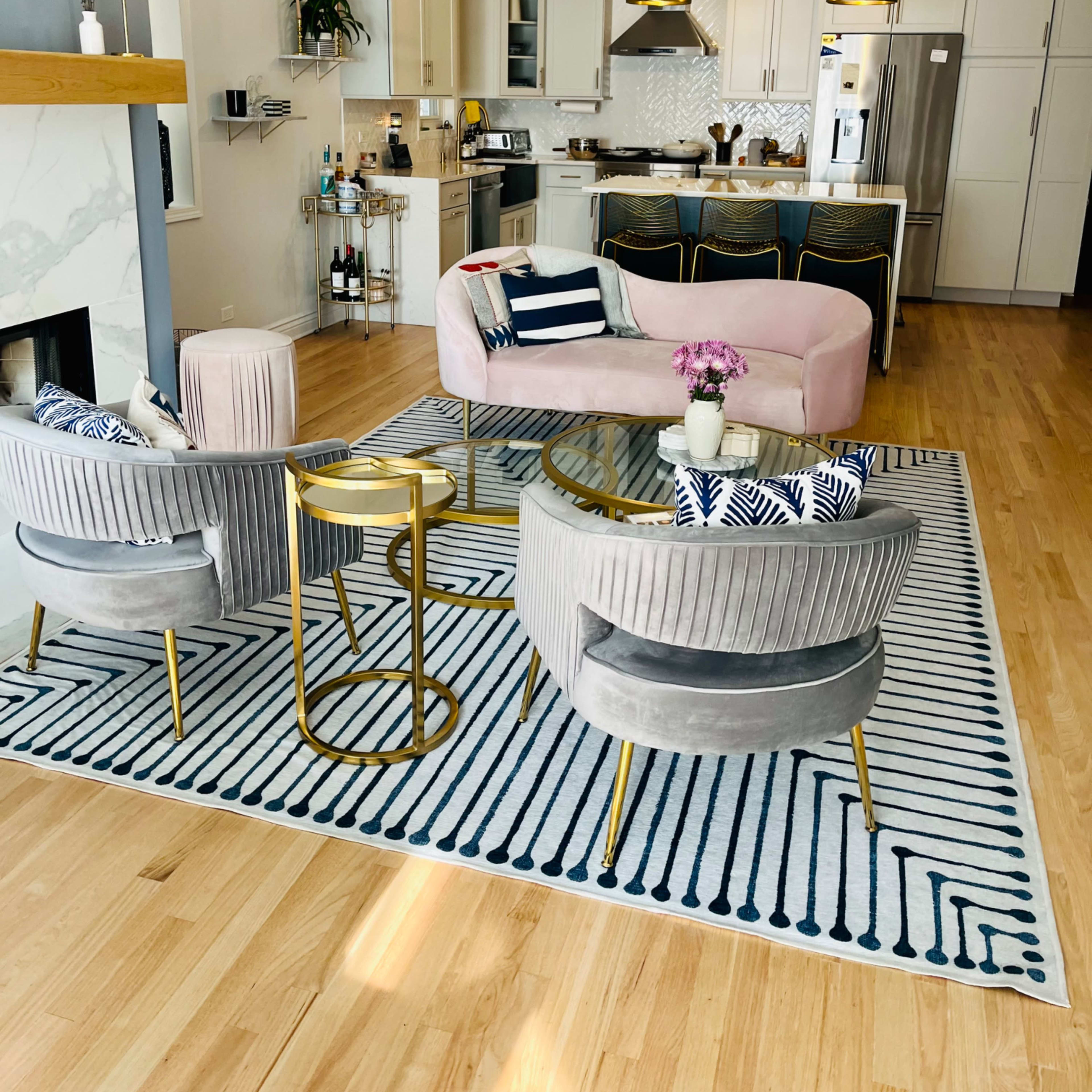 A modern living room with a pink sofa, two gray armchairs, a round gold side table, and a kitchen in the background.