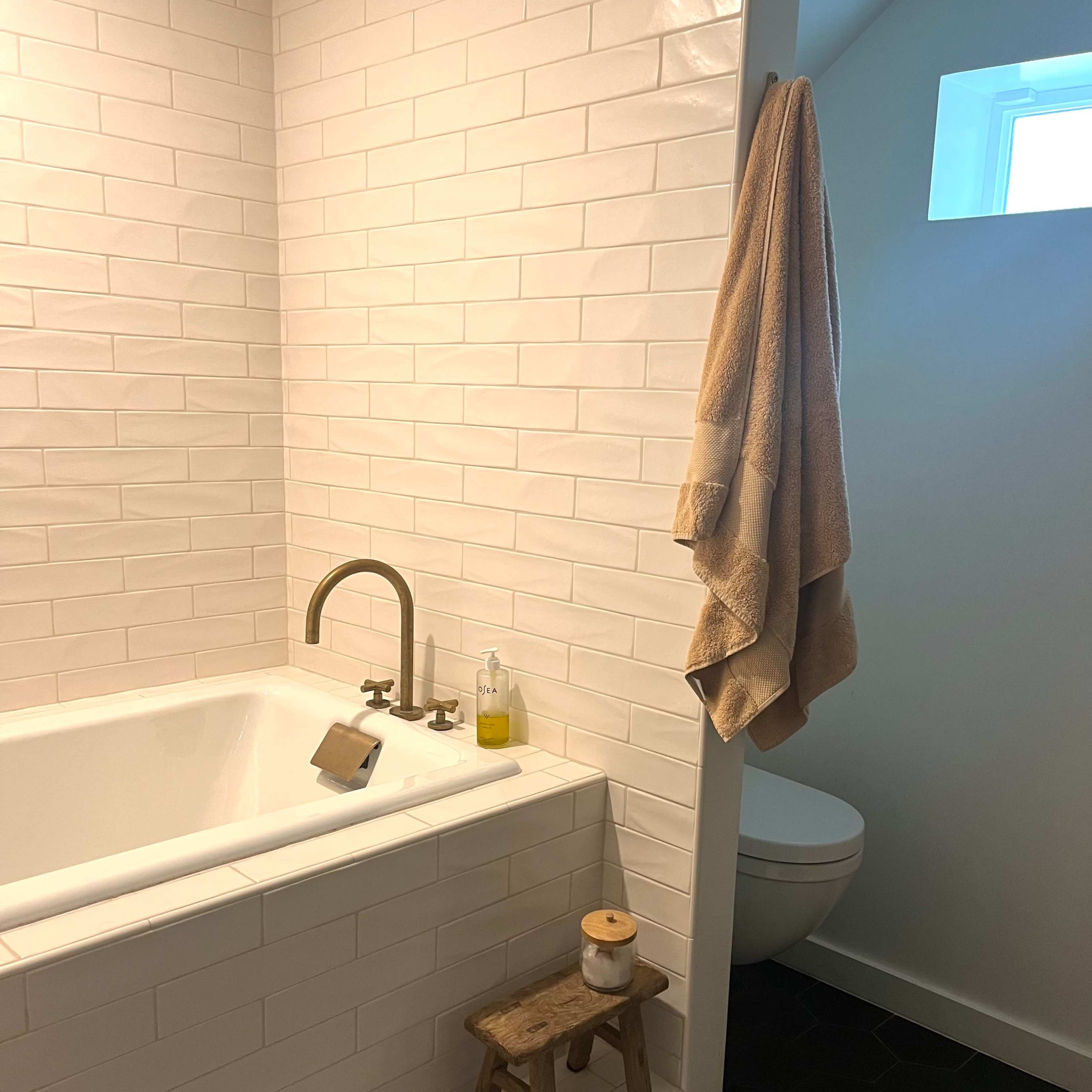 A modern bathroom featuring a white tiled bathtub, a small wooden stool with a candle, and a towel hanging on the wall, next to a toilet in a corner.
