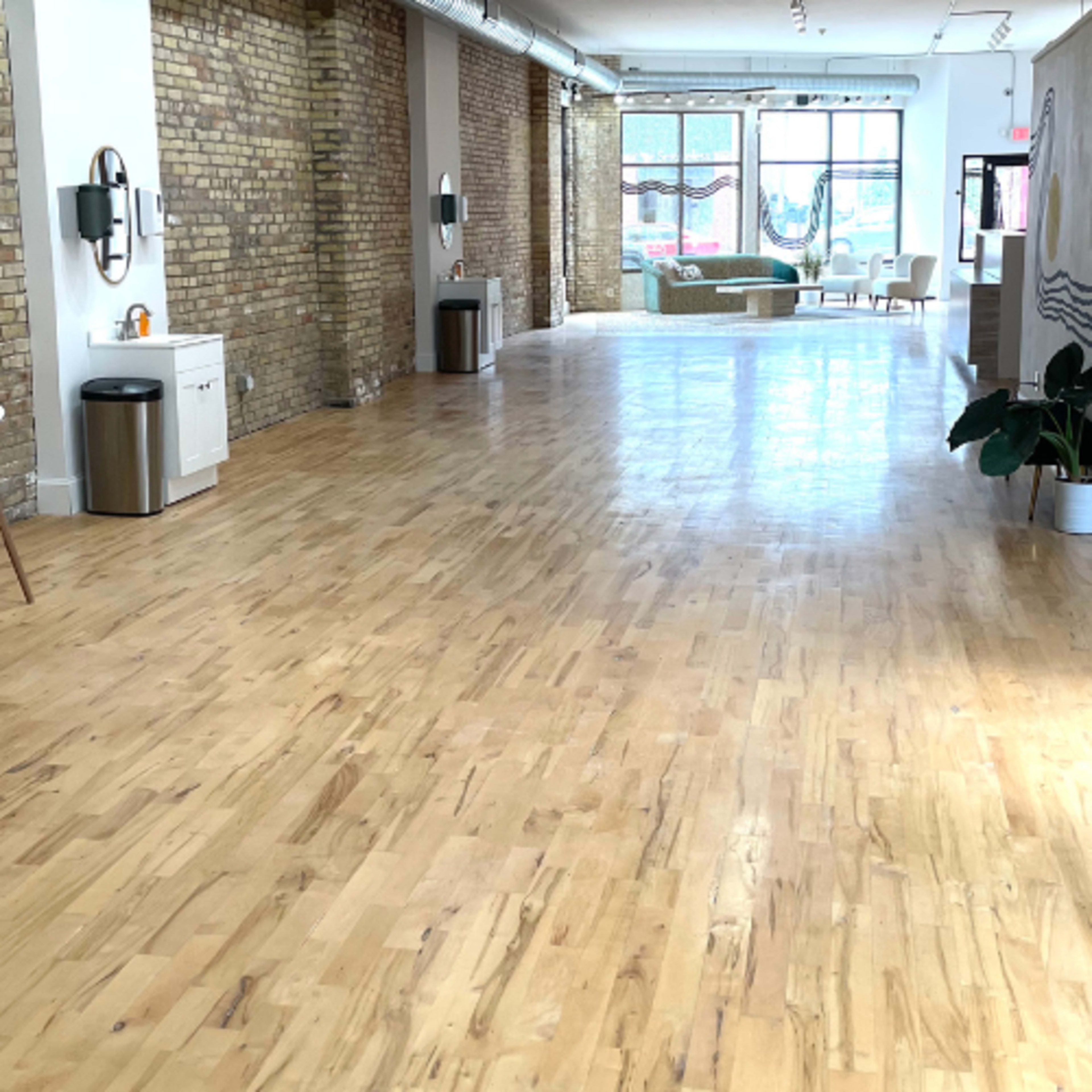 An empty, bright interior space with wooden flooring, exposed brick walls, and minimalist furniture.