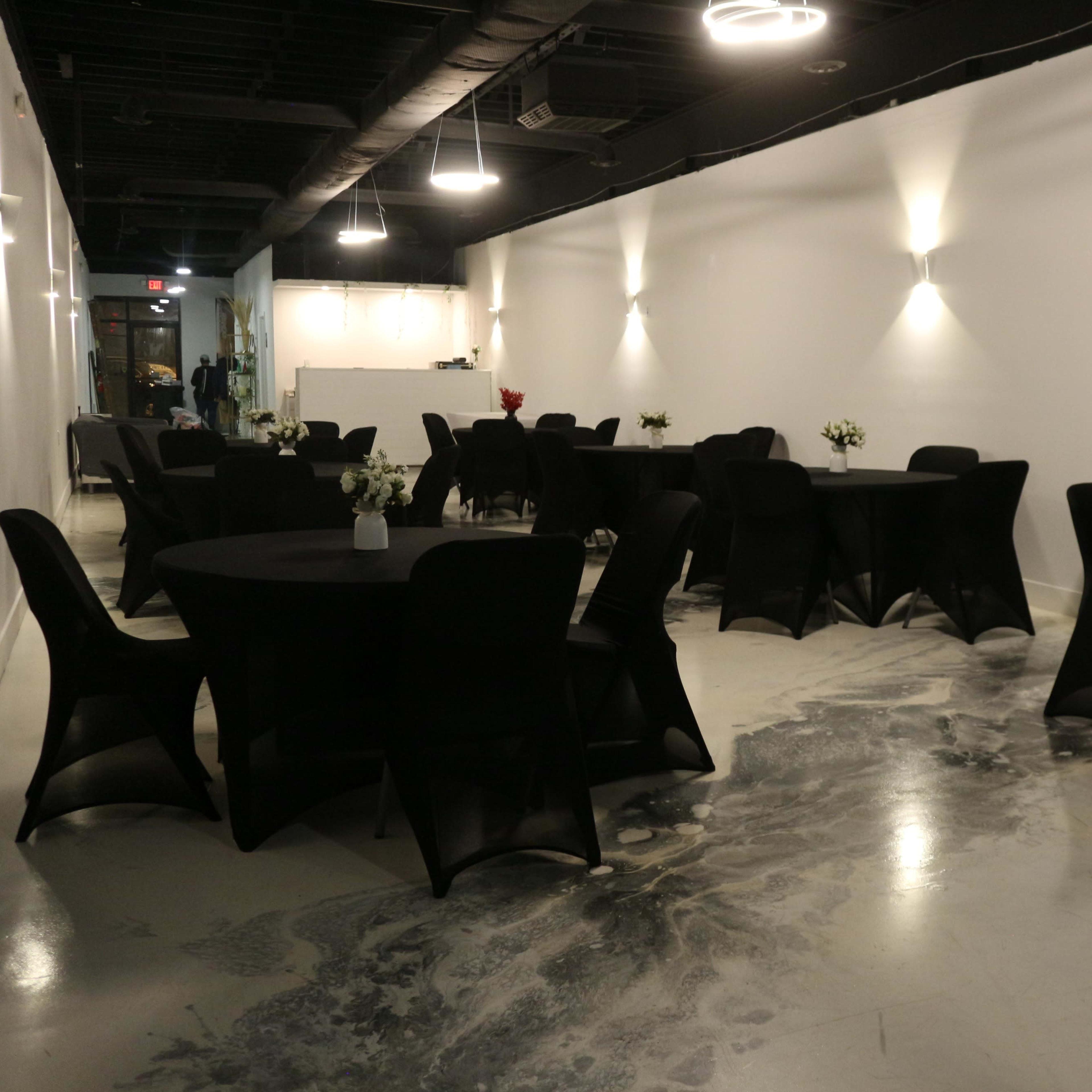 A spacious room with multiple round tables covered in black tablecloths, each adorned with small flower arrangements, set against a white wall and a polished concrete floor.