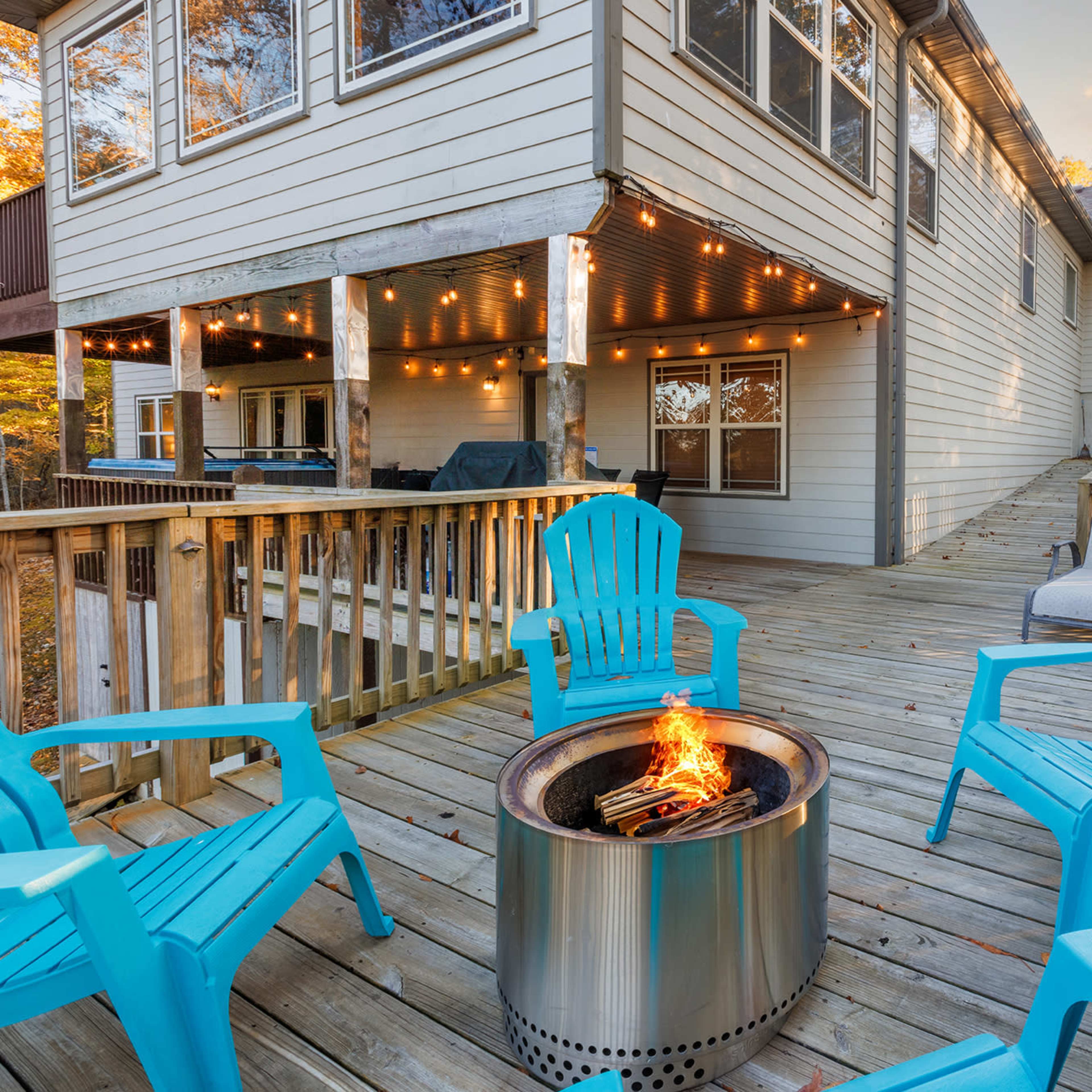 A wooden deck features a fire pit surrounded by blue Adirondack chairs, with a house in the background adorned with string lights.