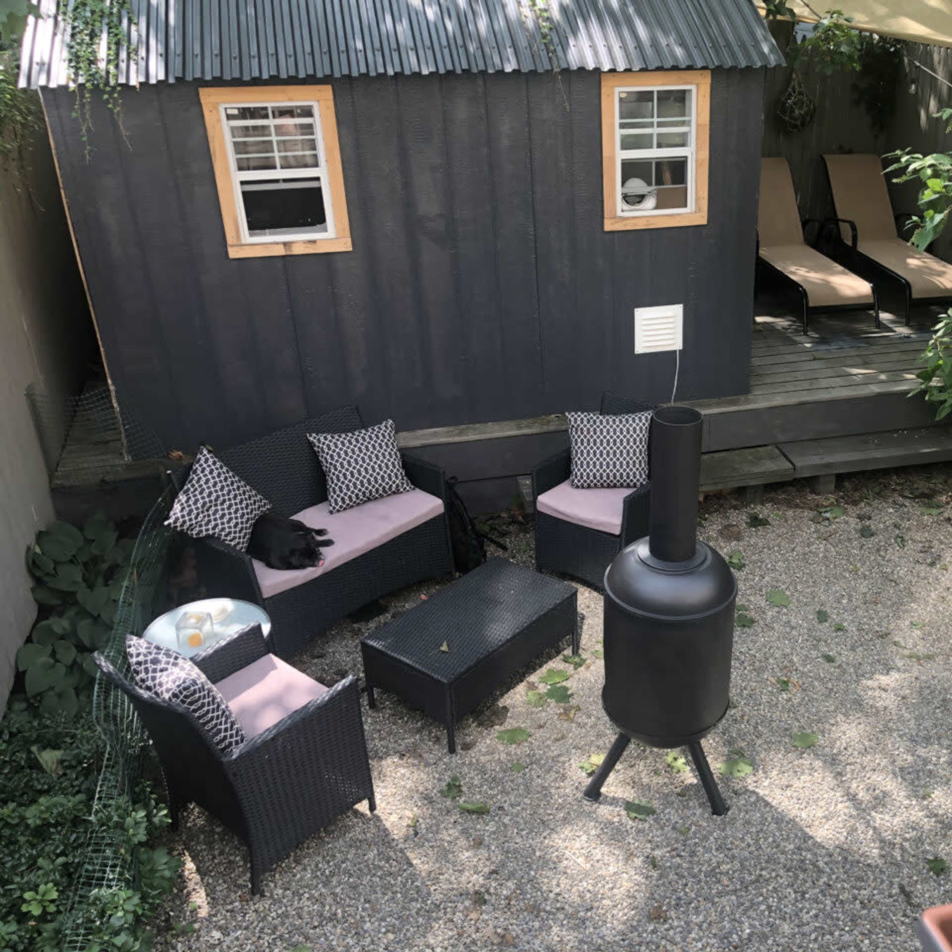 The scene shows a cozy outdoor space featuring a black shed, a gravel patio with seating, and a chimney-style fire pit surrounded by greenery.