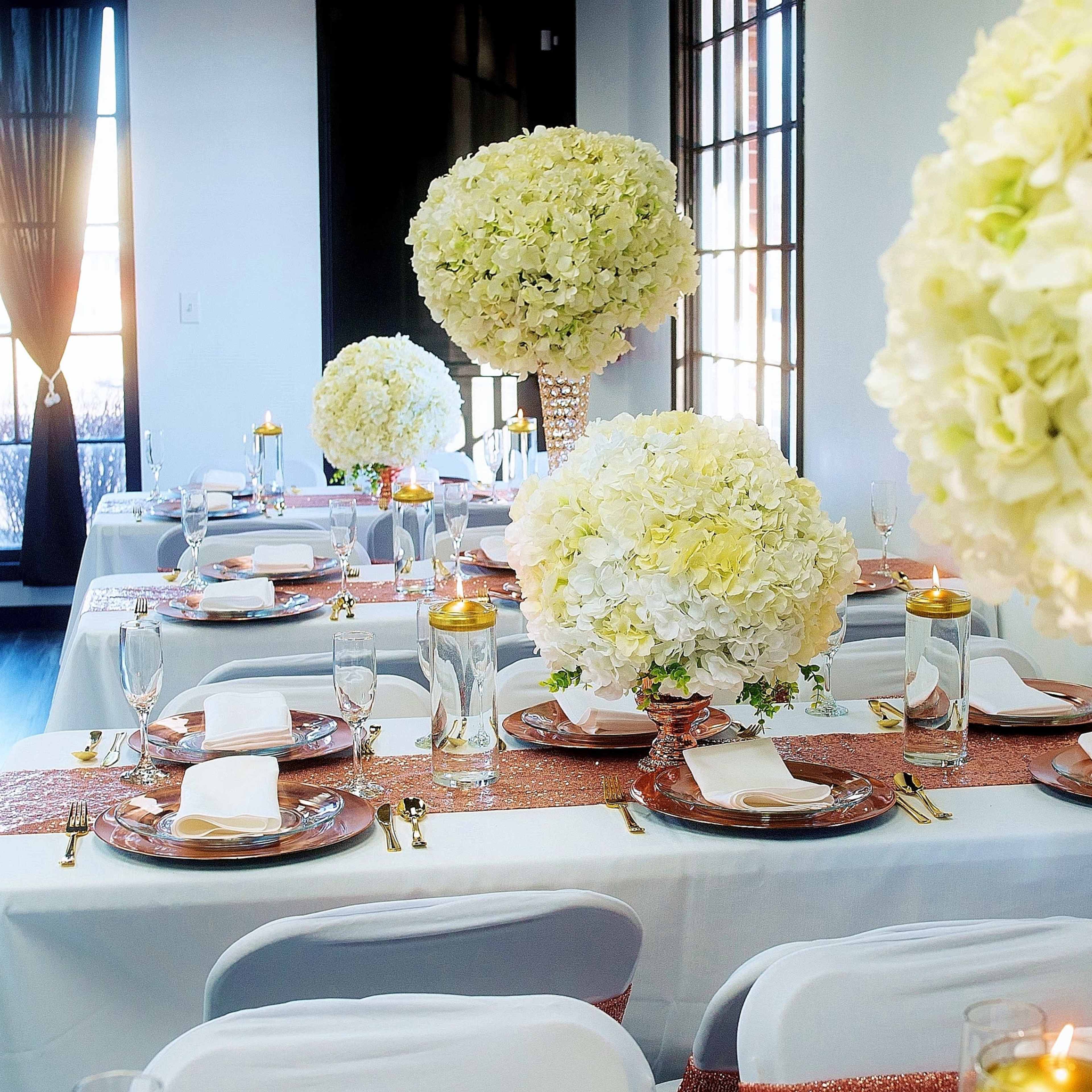 A elegantly set dining area with tables adorned with white floral centerpieces and sparkling table runners, illuminated by natural light through large windows.