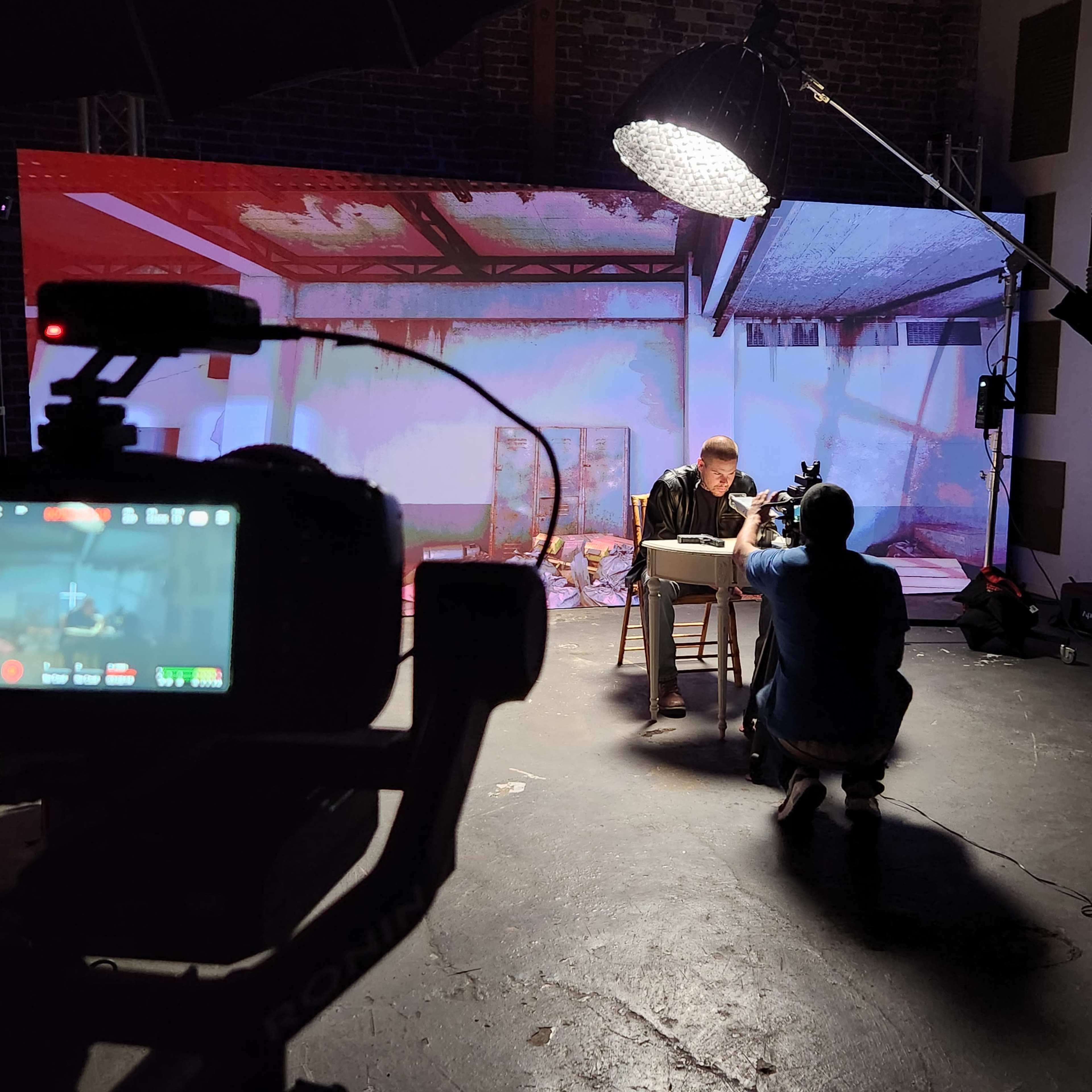 A video production setup shows a person sitting at a table while another person films them with a camera in a dimly lit industrial space.
