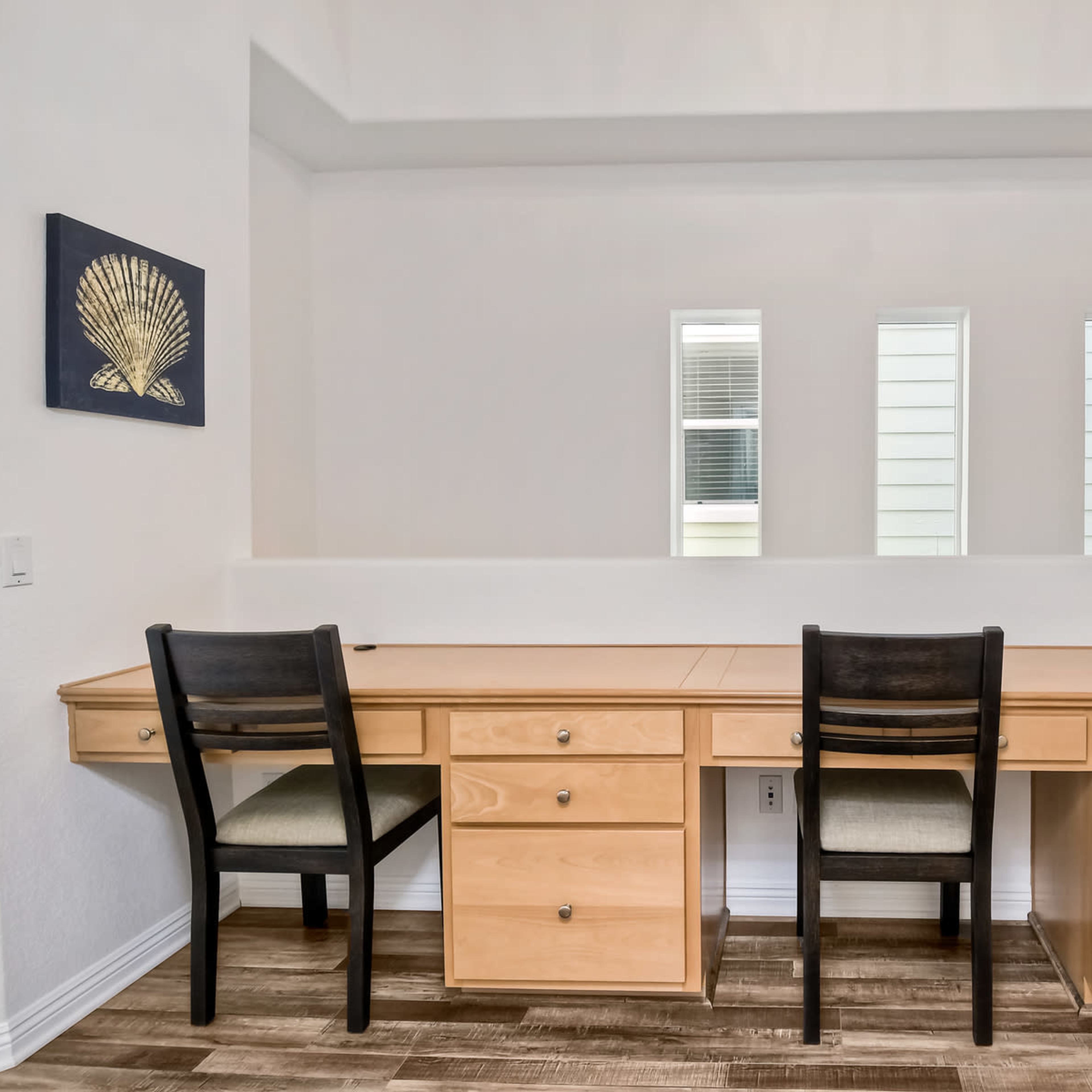 A wooden desk with two chairs is positioned against a white wall, featuring three vertical windows and a shell-themed artwork on the left.