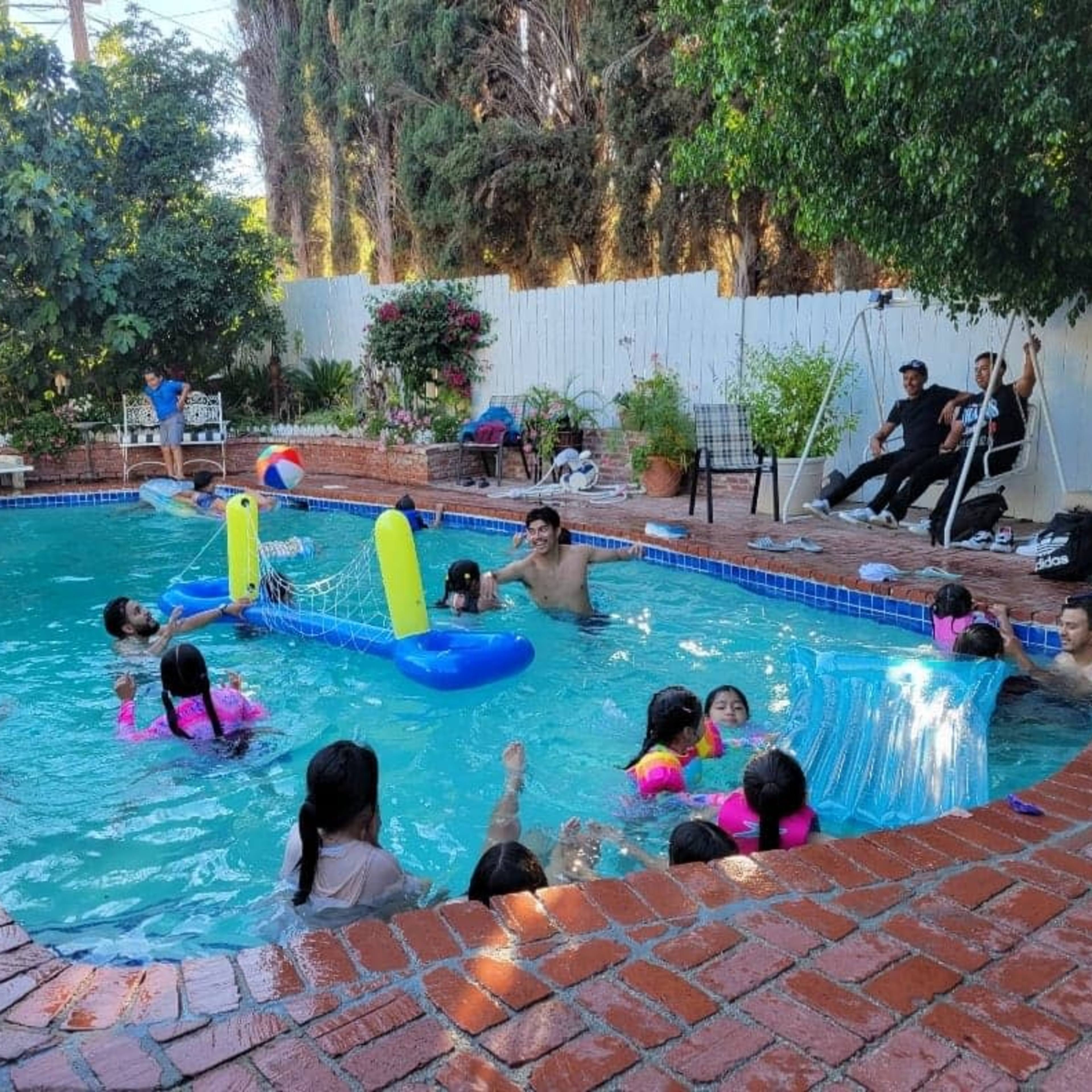 A group of children and adults are gathered around a residential pool filled with inflatable toys, enjoying the warm weather.