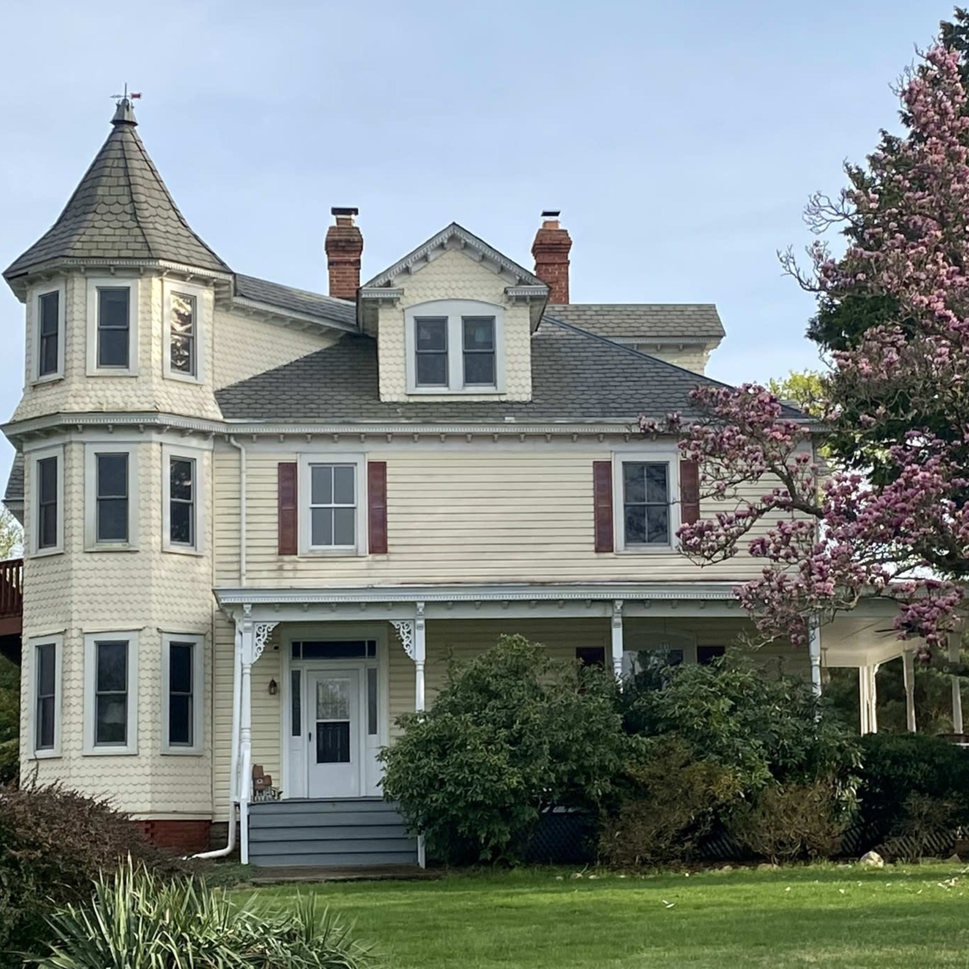 A large, Victorian-style house with a turret and a flowering tree in the front yard.