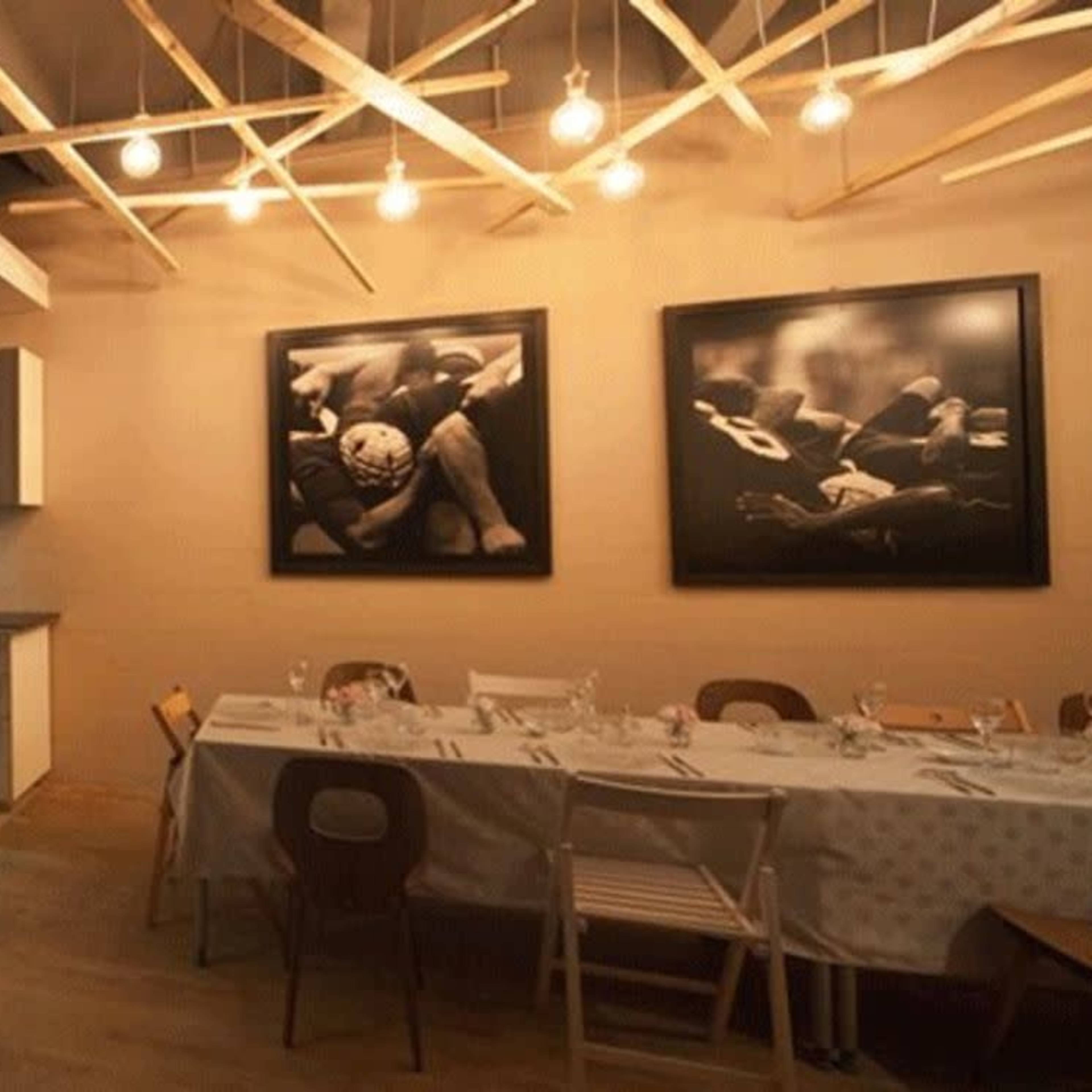 A modern kitchen area connected to a dining space with a long table set for a meal, illuminated by pendant lights and framed black and white photographs on the walls.