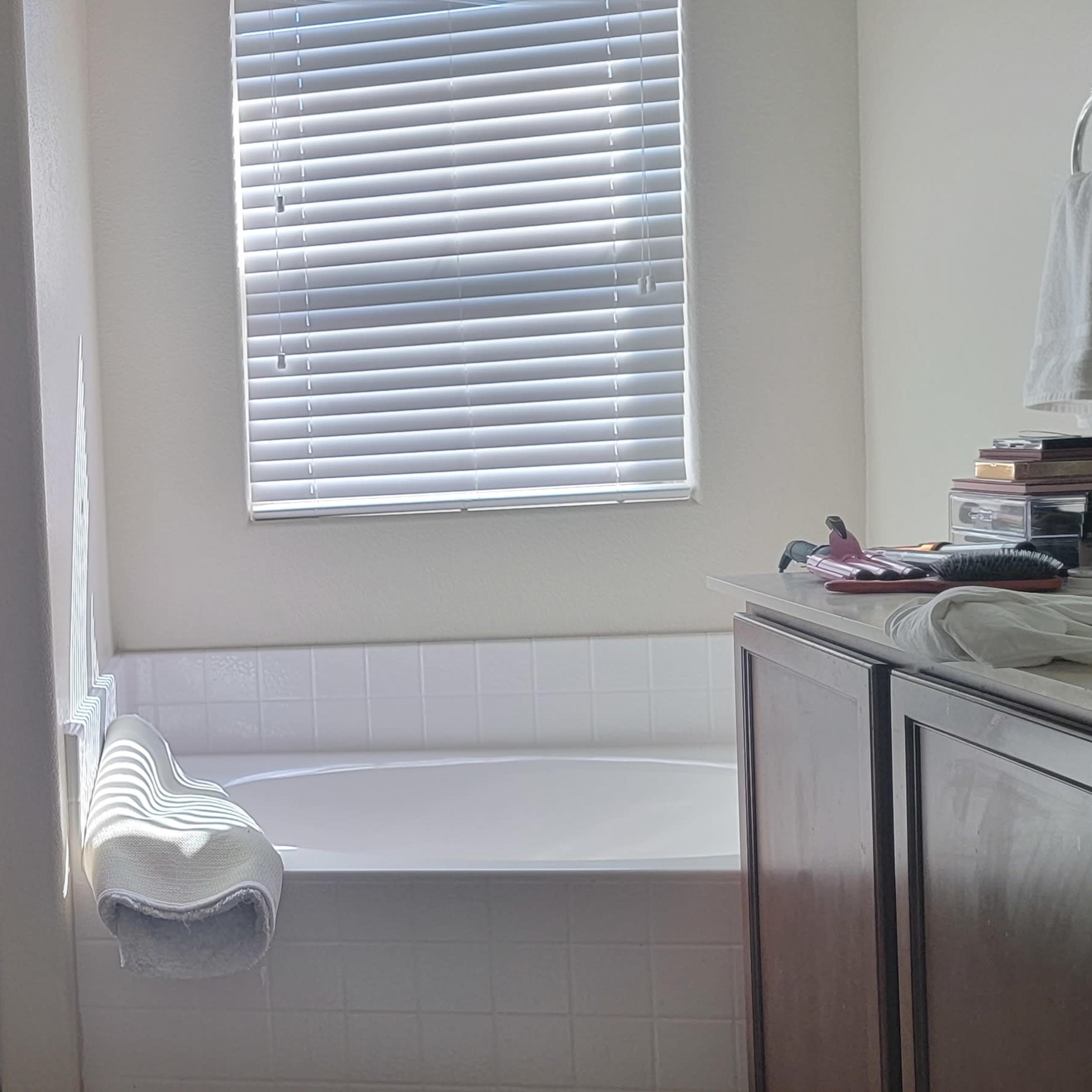 A bathroom features a glass shower stall beside a bathtub, with towels hanging on a hook and a window with blinds.