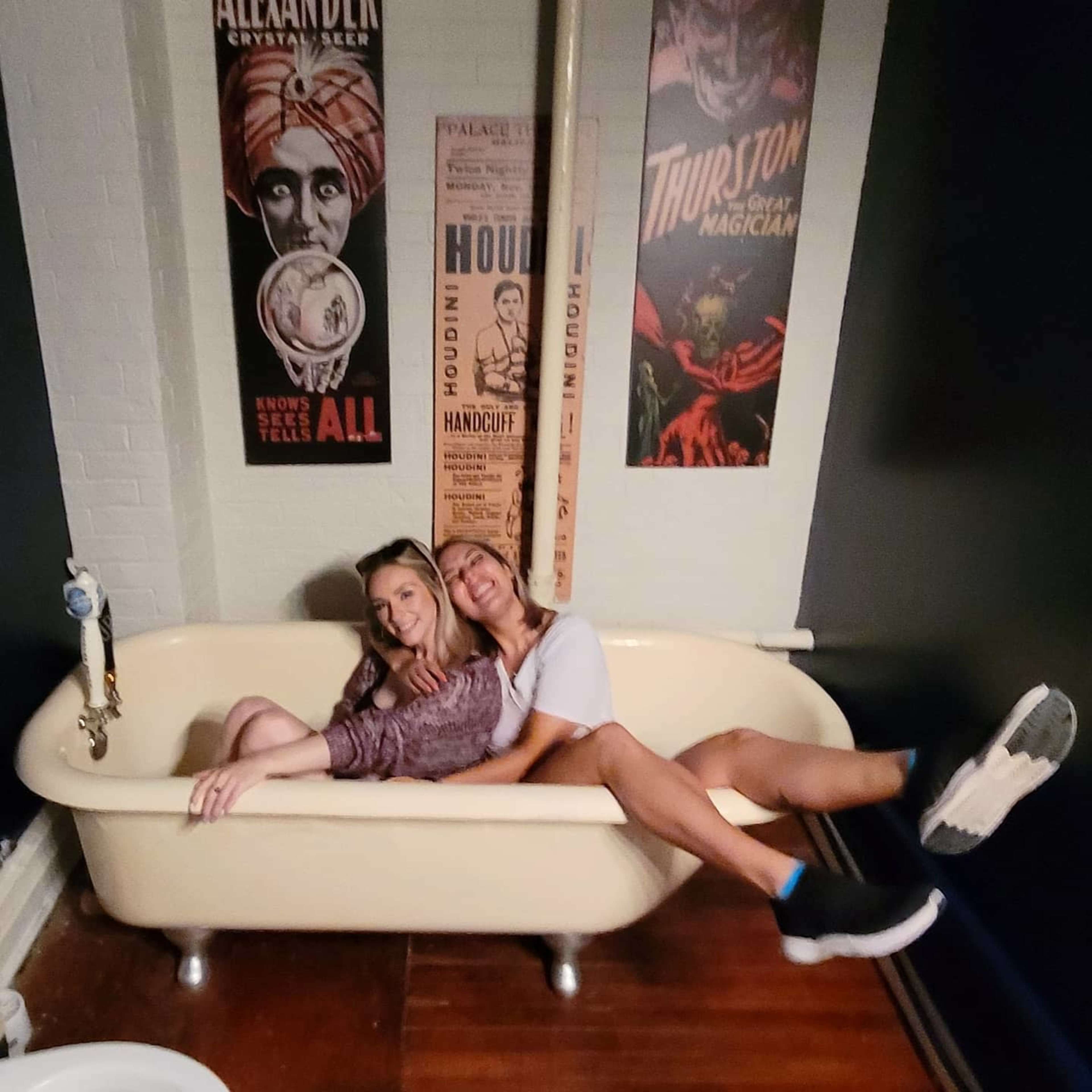 Two women sit together in a vintage bathtub surrounded by theatrical posters on the walls.