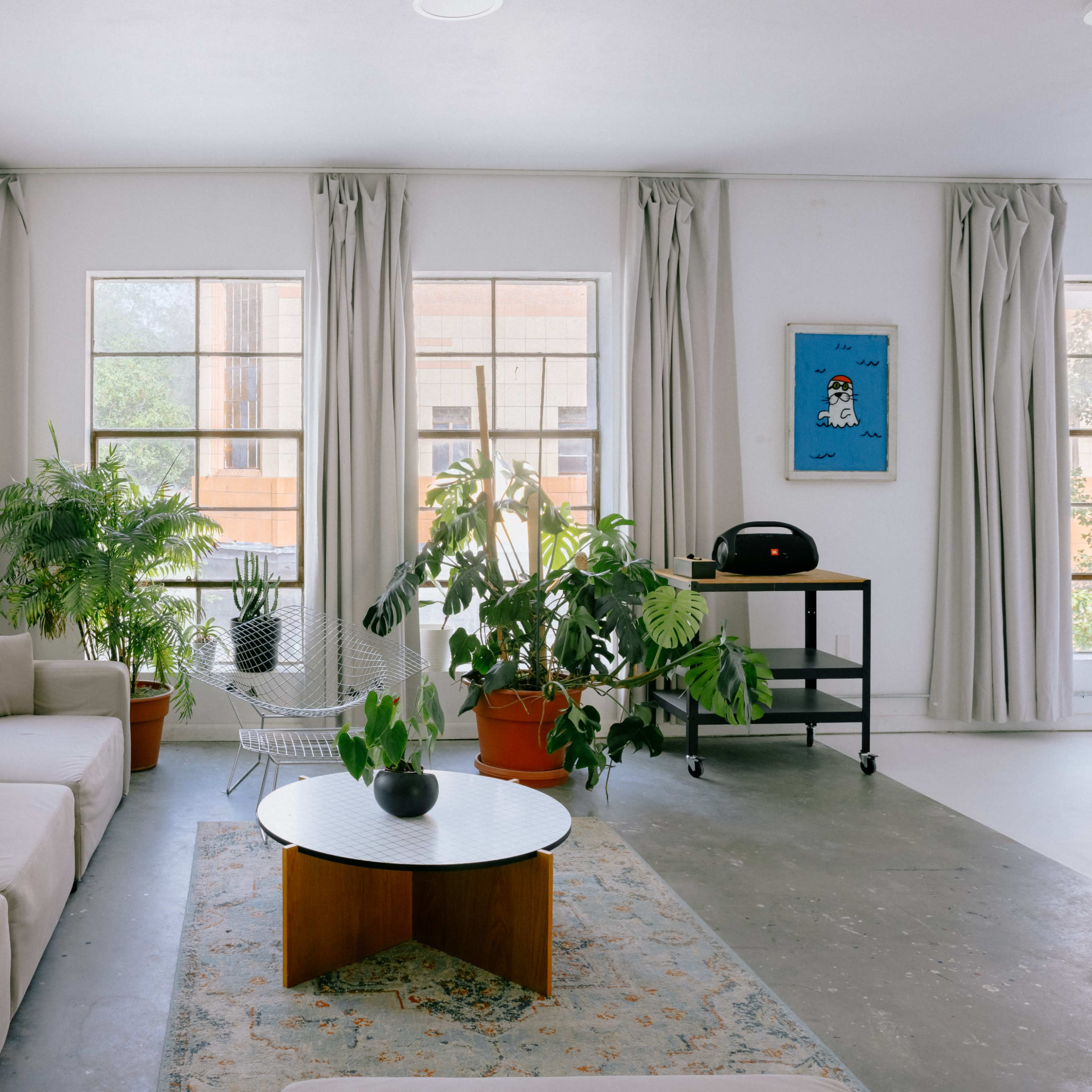 A bright, minimalist living space with a light-colored sofa, a round coffee table, and various potted plants, illuminated by natural light through large windows.