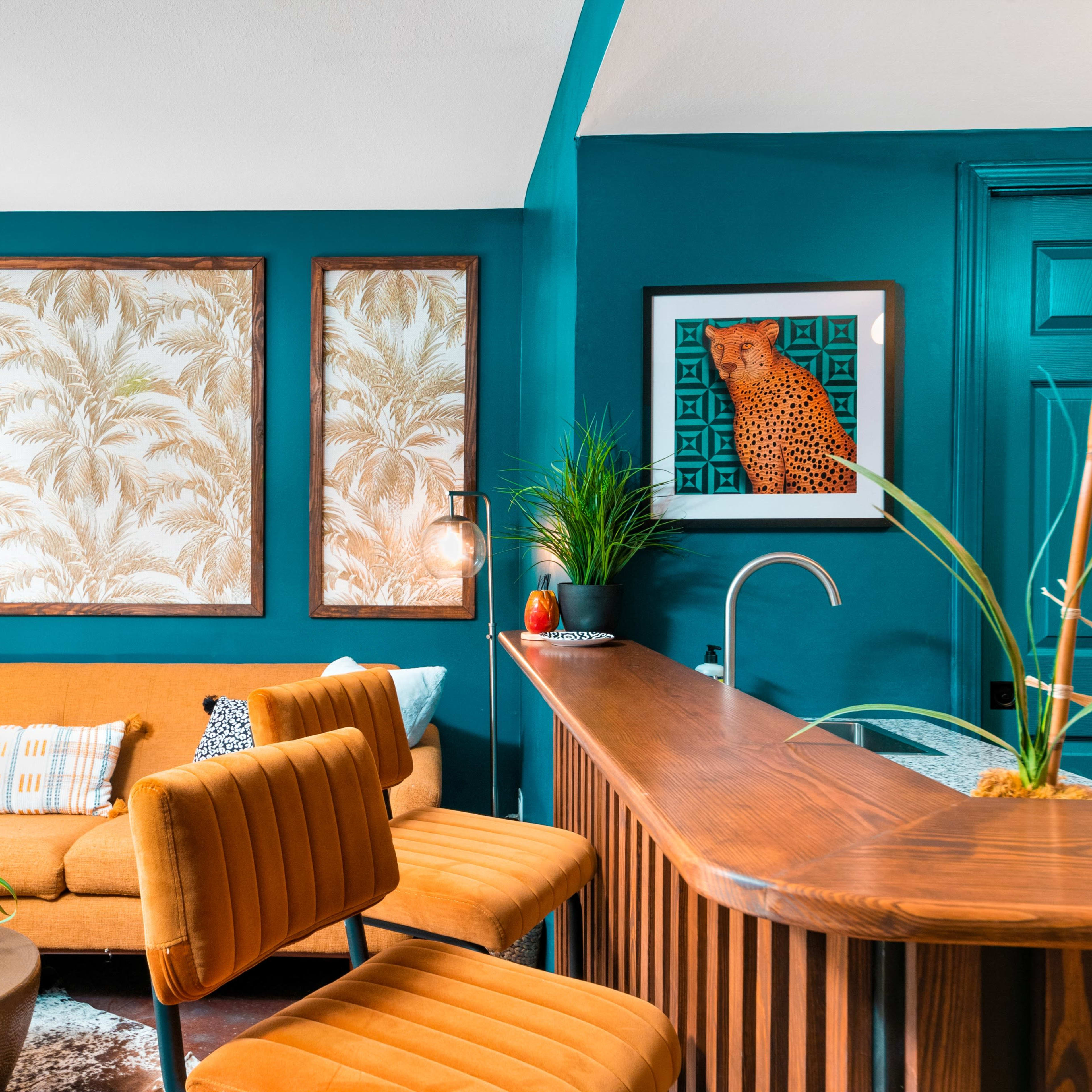 A bright living room featuring a mustard-colored sectional sofa, framed botanical prints on the turquoise walls, and a sleek bar counter with modern seating.