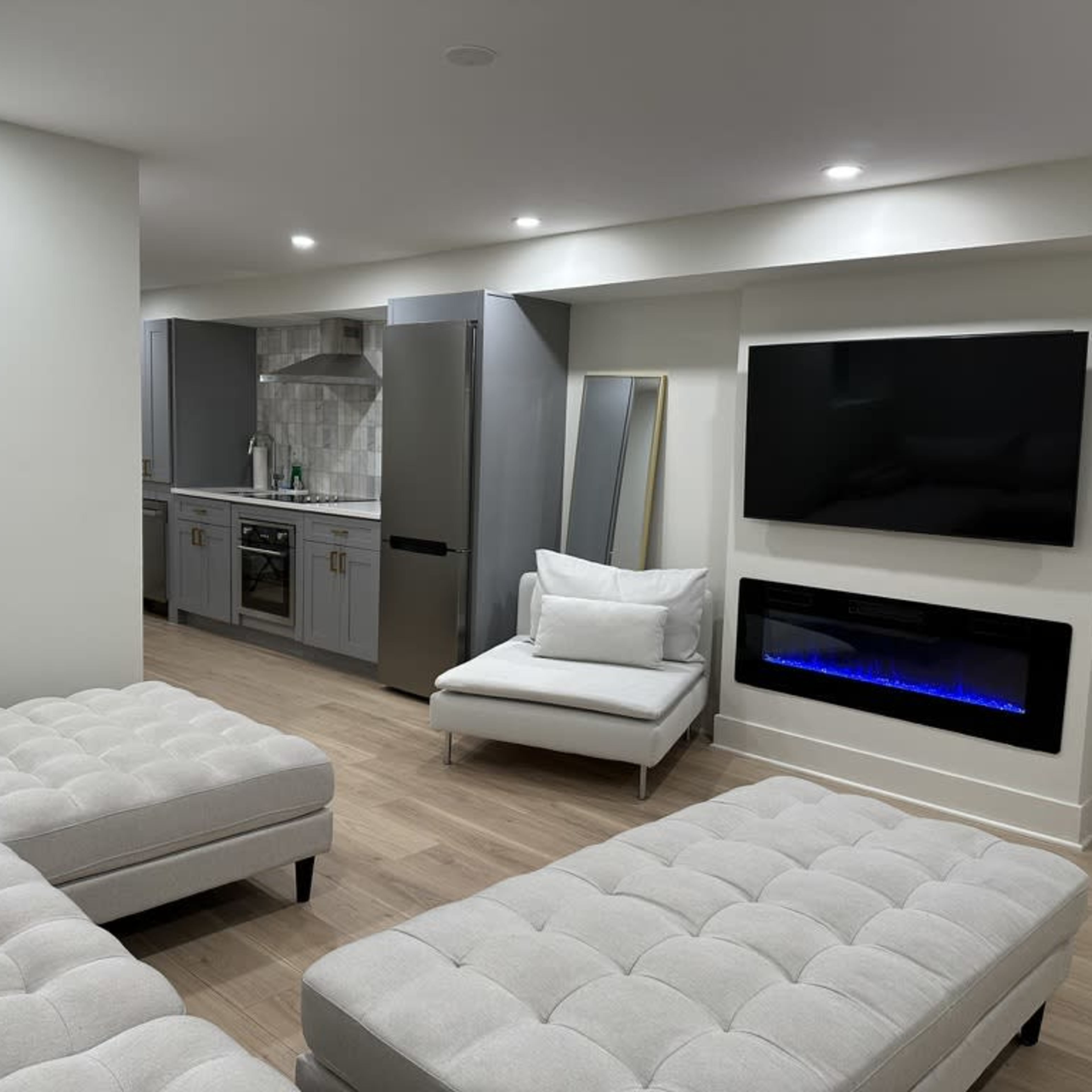 A modern living room with light-colored furniture, a fireplace, a large wall-mounted television, and a kitchen area visible in the background.