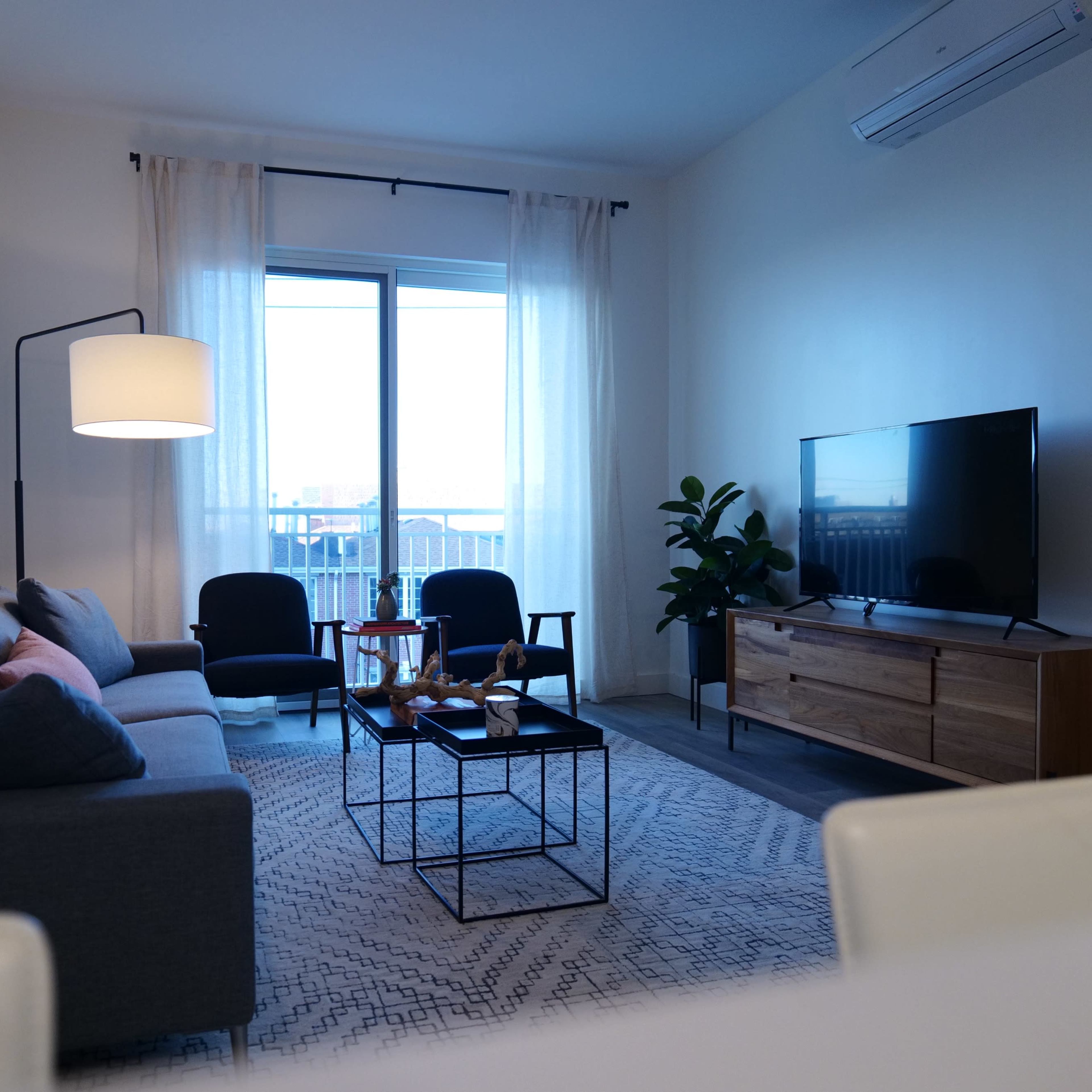 The living room features a gray sofa, two black chairs, a wooden TV stand, and a large window with sheer curtains.