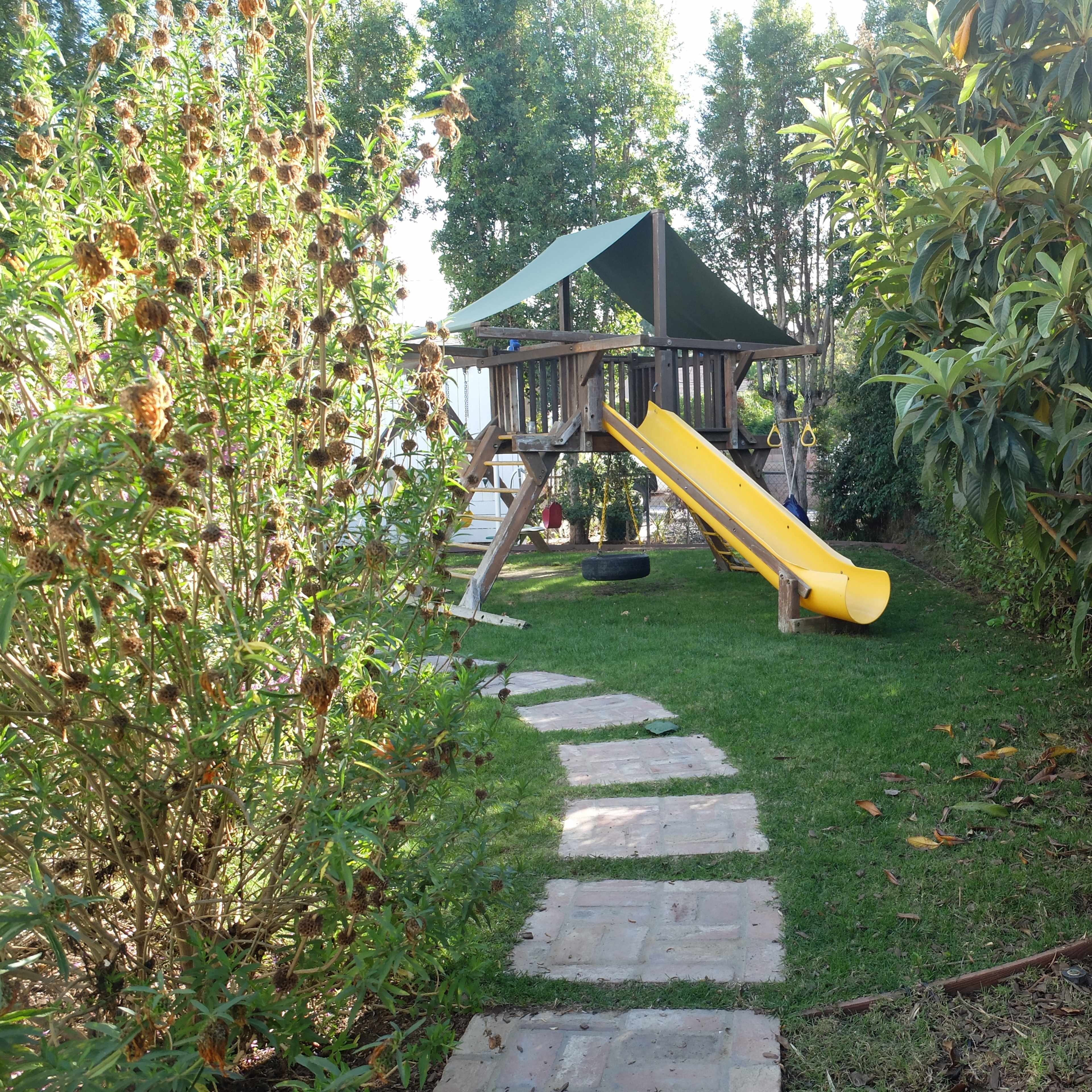 A playset with a yellow slide and a canopy is situated in a grassy area surrounded by bushes and a stone pathway.