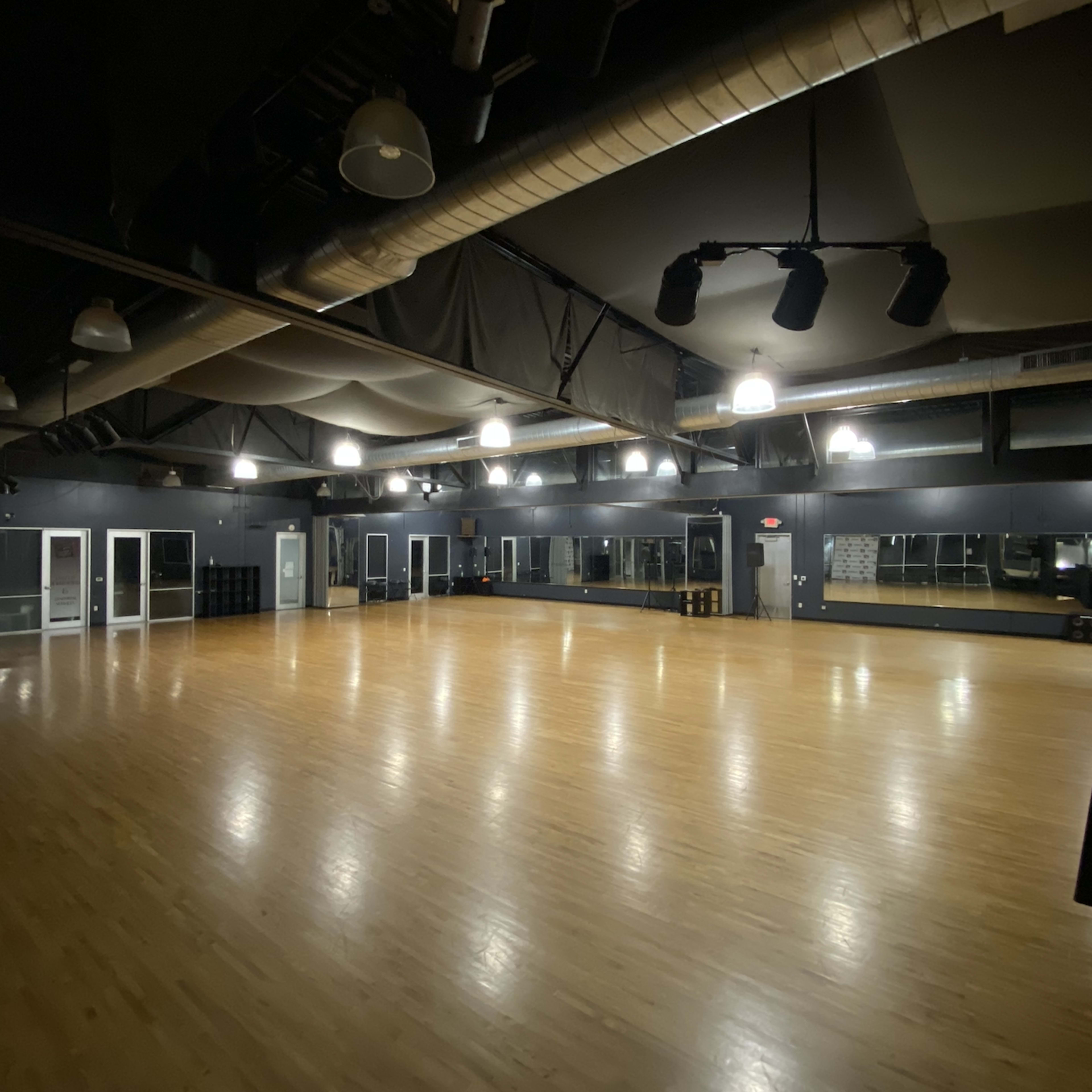 A large, empty dance studio with wooden flooring, mirrors on one wall, and overhead lighting in a dark-colored space.