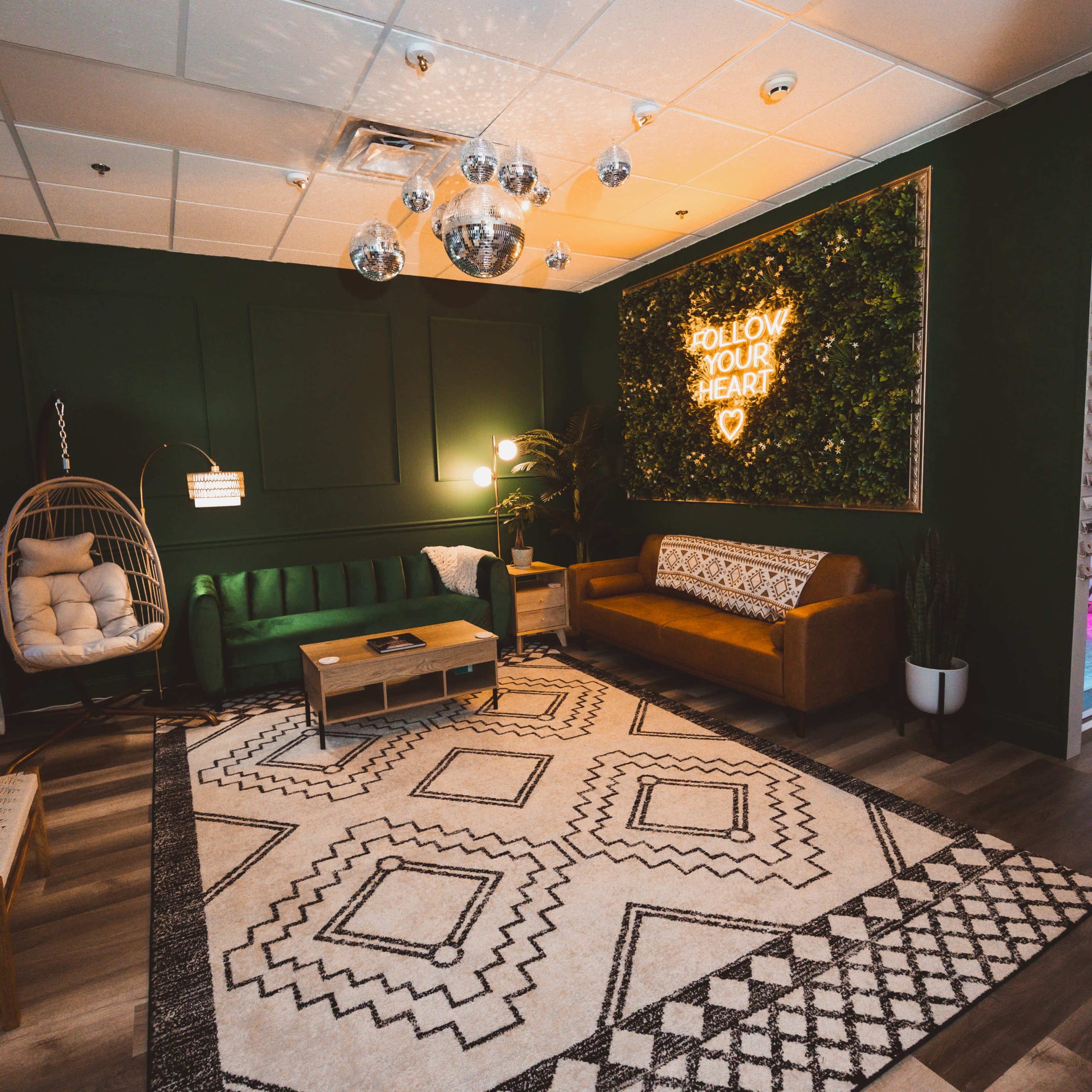 A modern lounge area featuring green walls, a hanging chair, two sofas, a coffee table, and decorative plants with a neon sign.