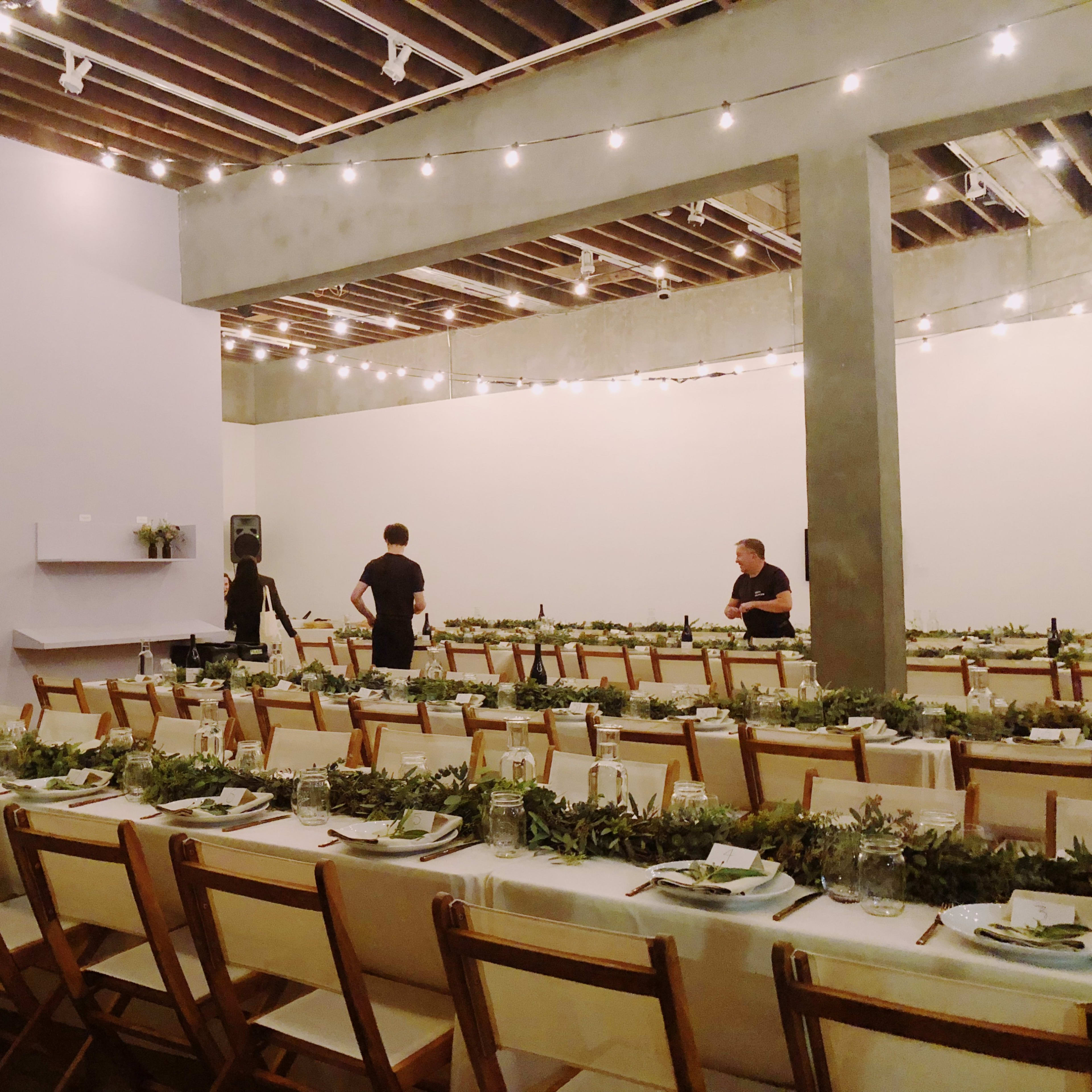A spacious dining area is set with long tables adorned with greenery and table settings, while two people prepare the space under string lights.