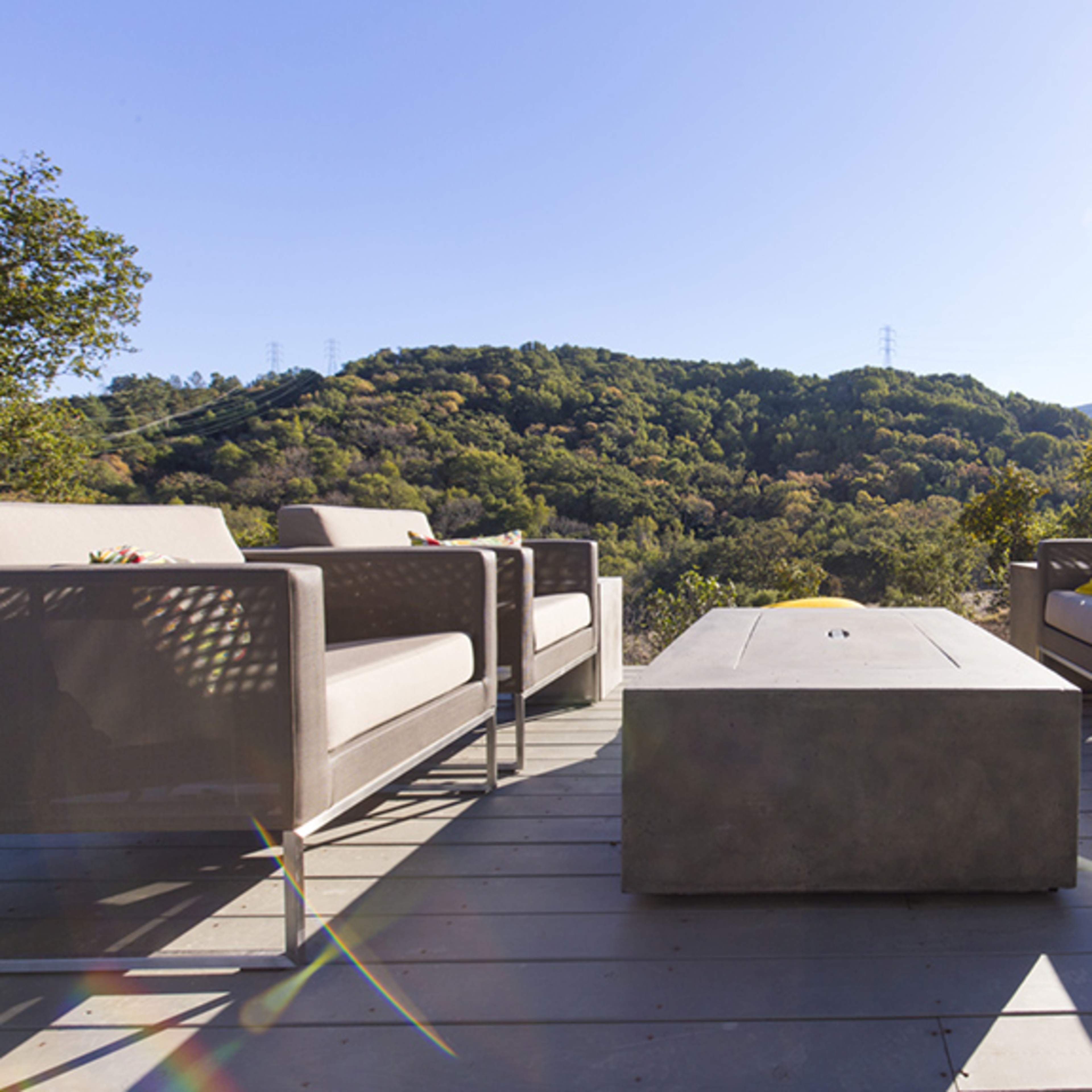 A modern outdoor seating area features two sofas and a rectangular coffee table, surrounded by lush green hills and clear blue skies.