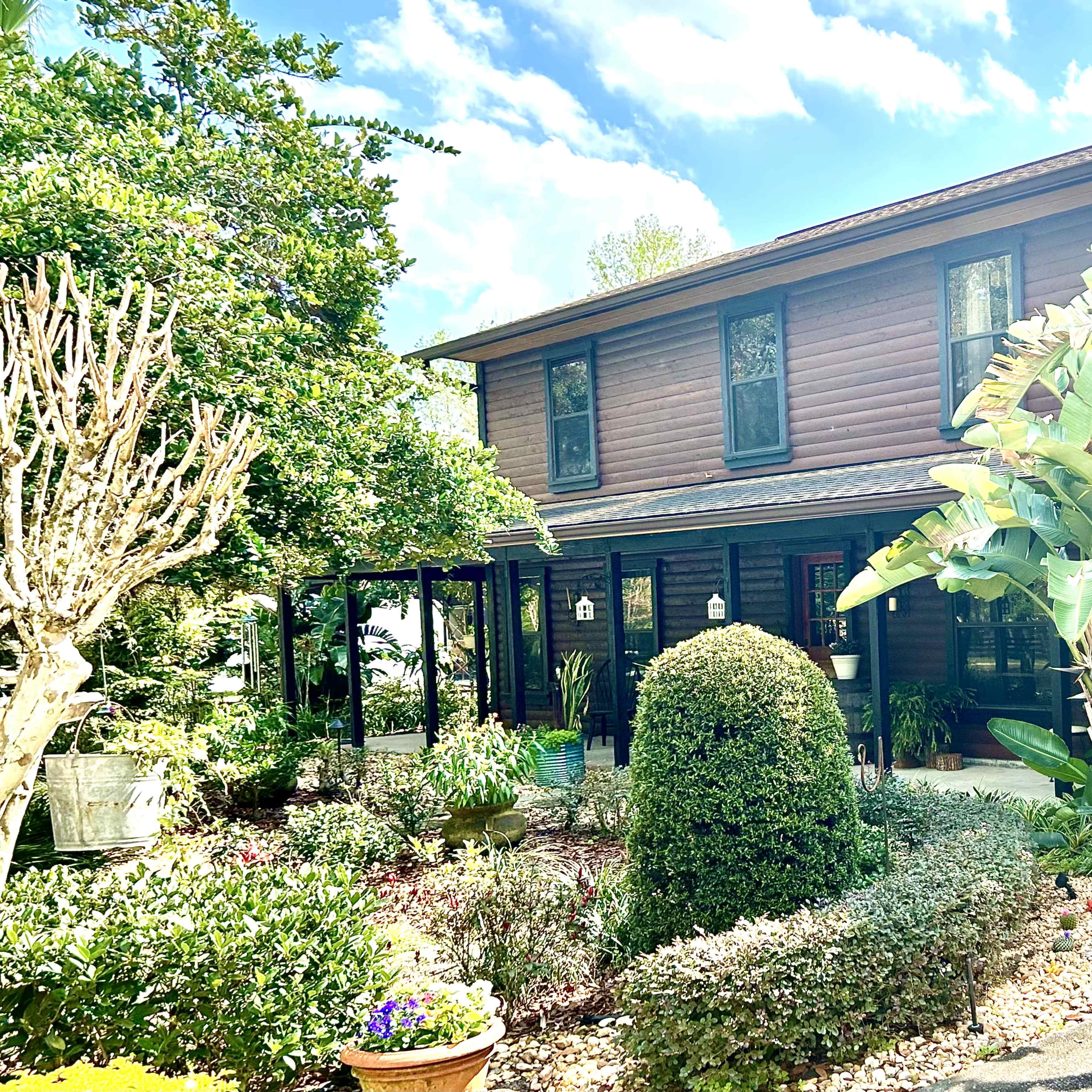 A two-story wooden house with a covered porch and lush landscaping featuring various plants and shrubs.