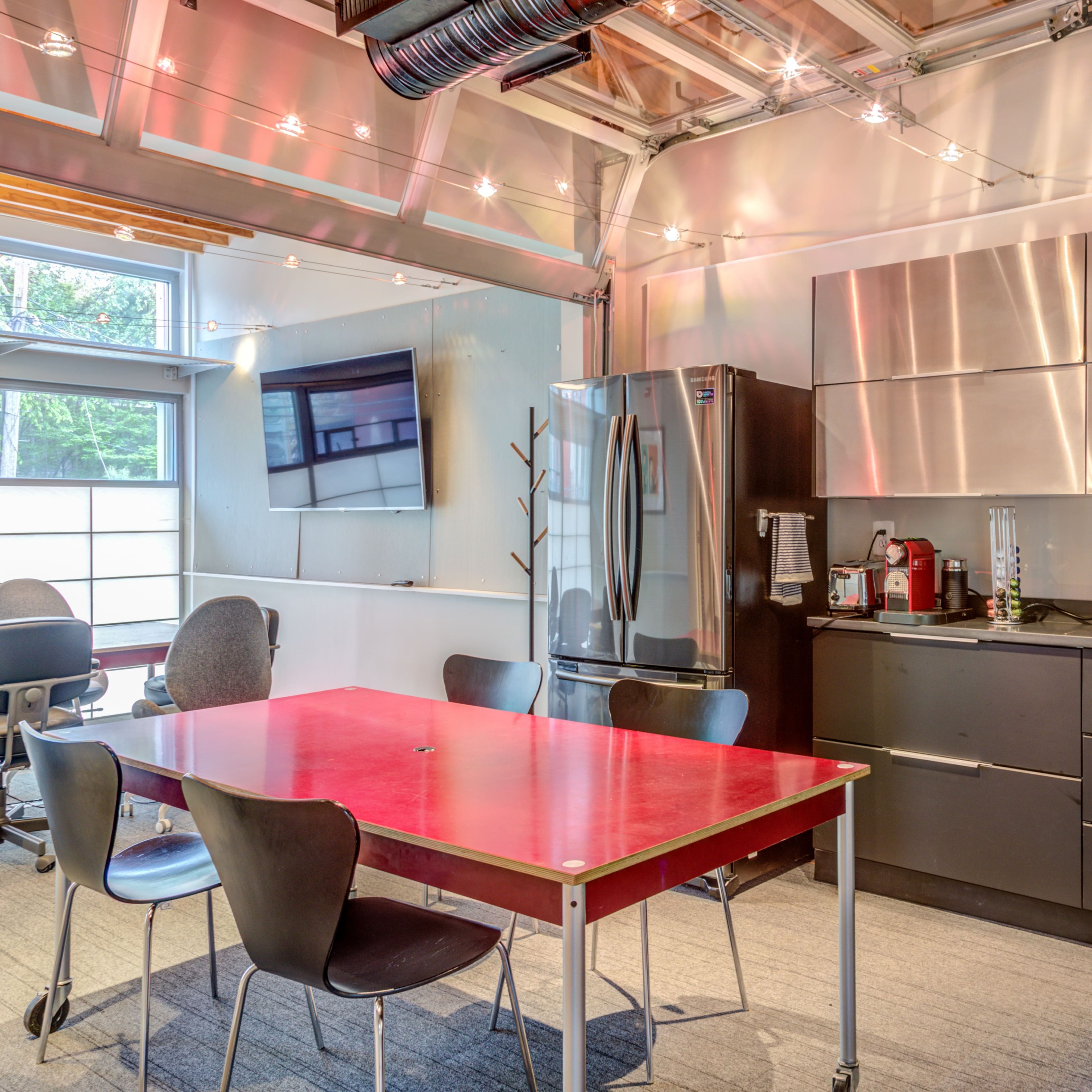 A modern office kitchen with a red table, black cabinets, stainless steel appliances, and seating arranged nearby.