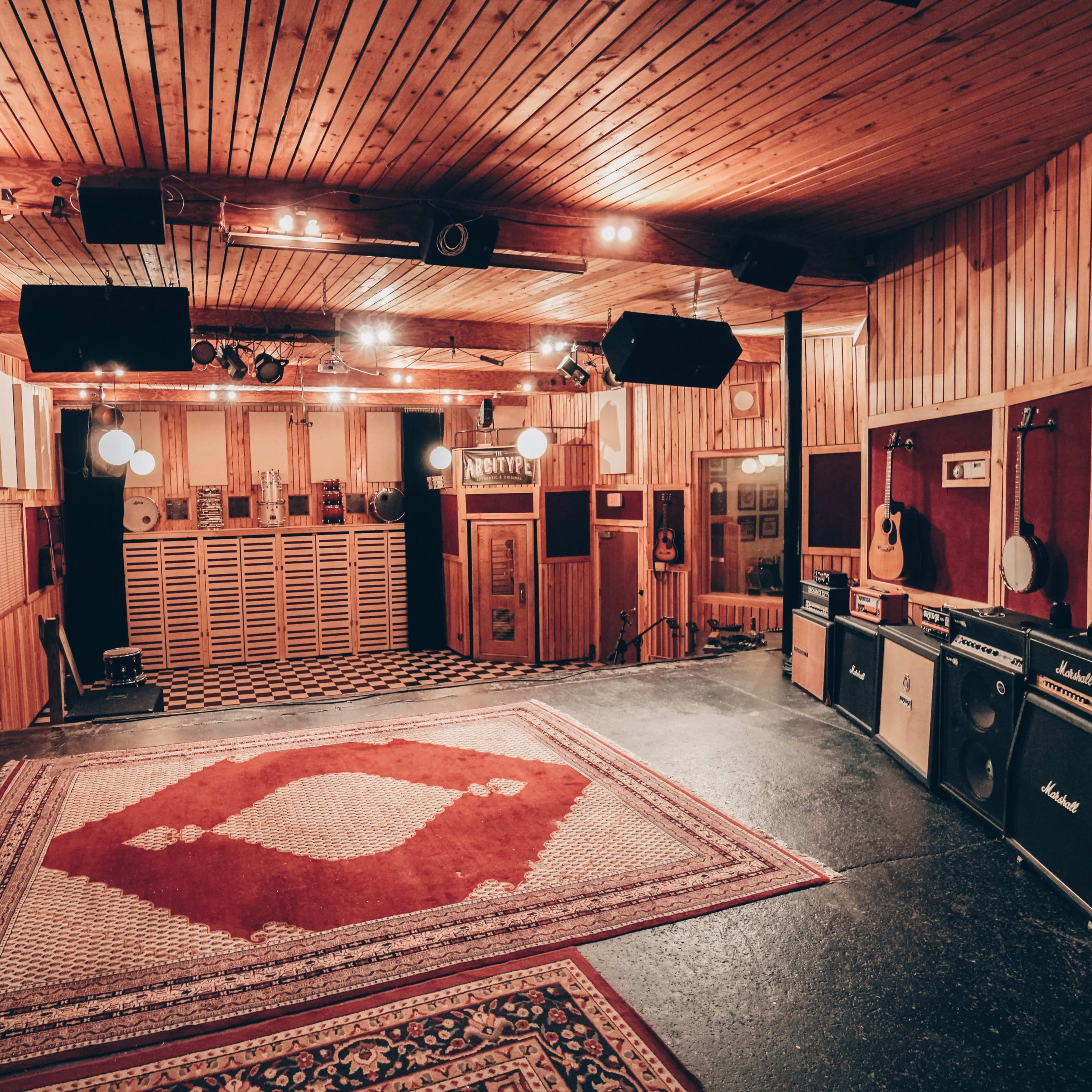 A spacious recording studio with wooden walls, a patterned rug on the floor, and musical instruments including guitars and amplifiers positioned around the room.