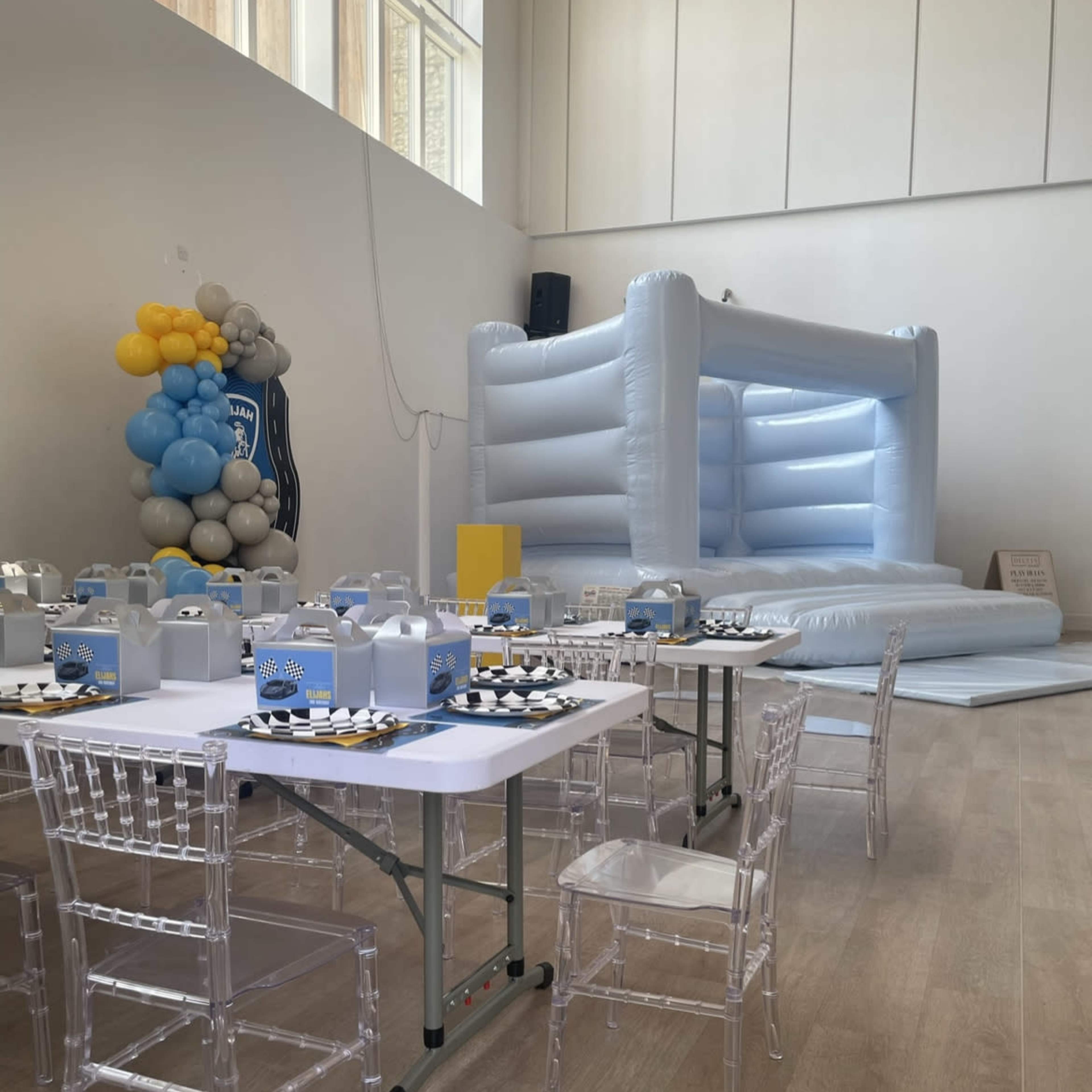 A spacious indoor venue set up for a celebration, featuring a large inflatable bounce house and several tables with clear chairs arranged around them.