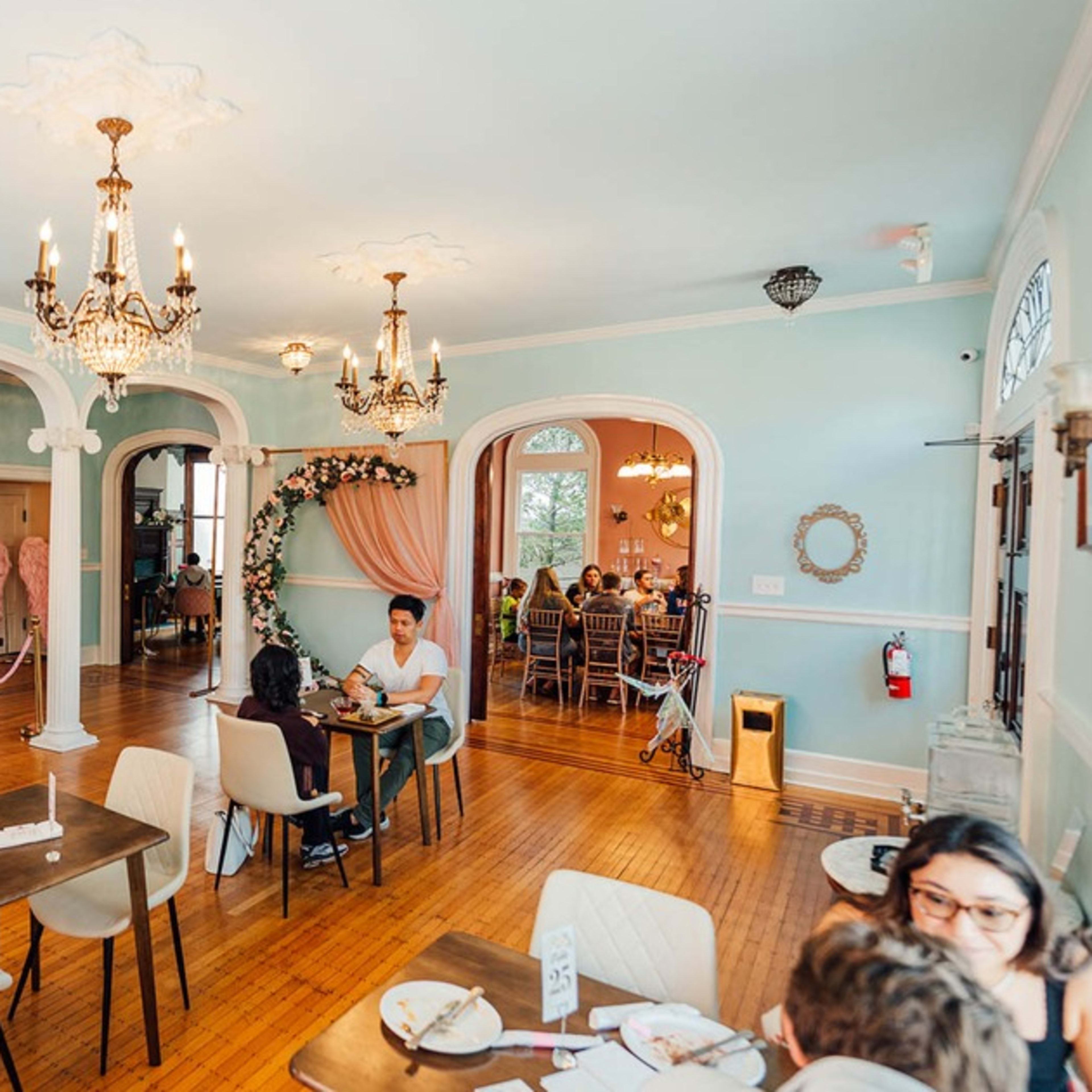 A spacious, pastel-colored café featuring elegant chandeliers, a staircase, and various seating options.