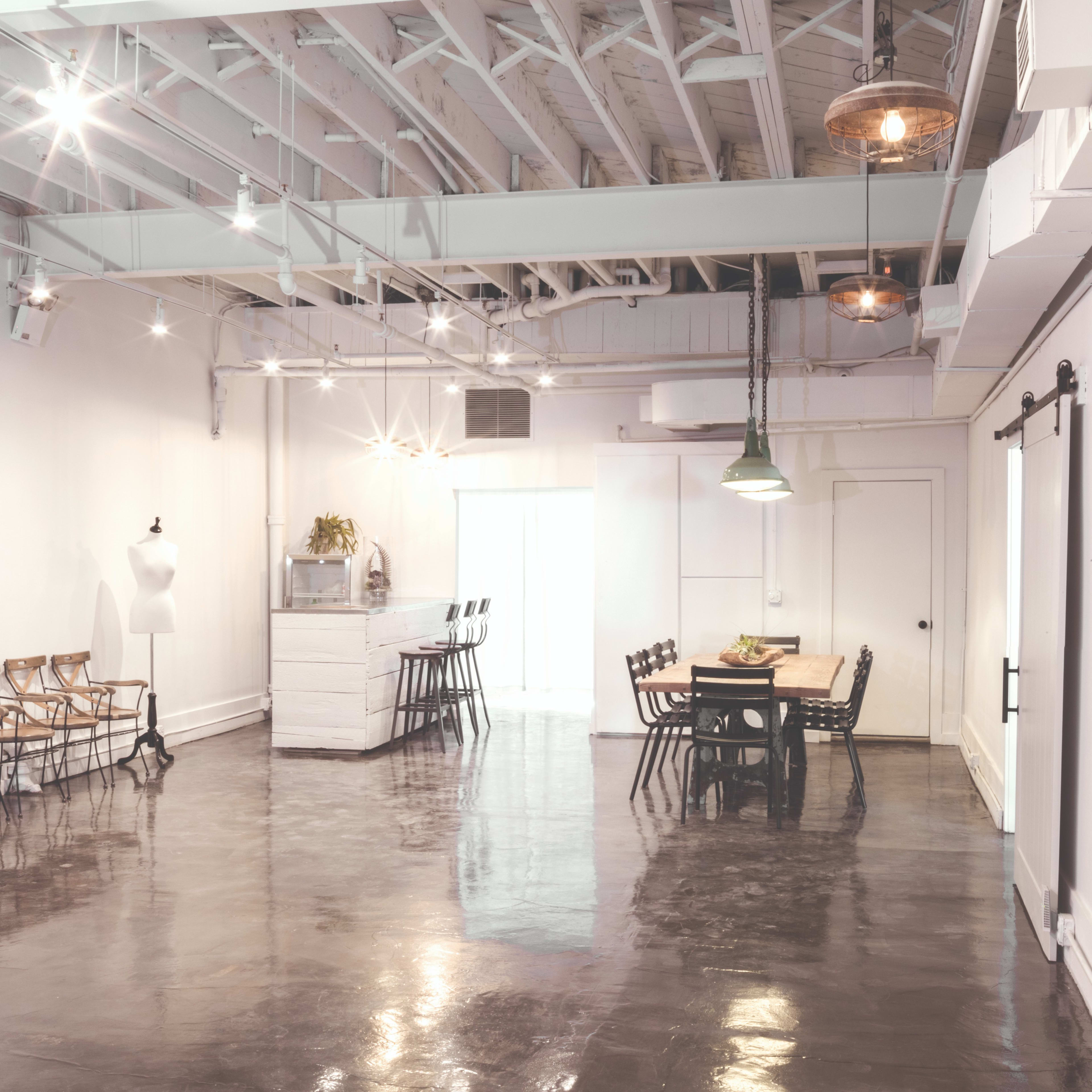 A spacious, well-lit room with a polished concrete floor, featuring a dining table and several chairs, along with a mannequin and a white dresser.