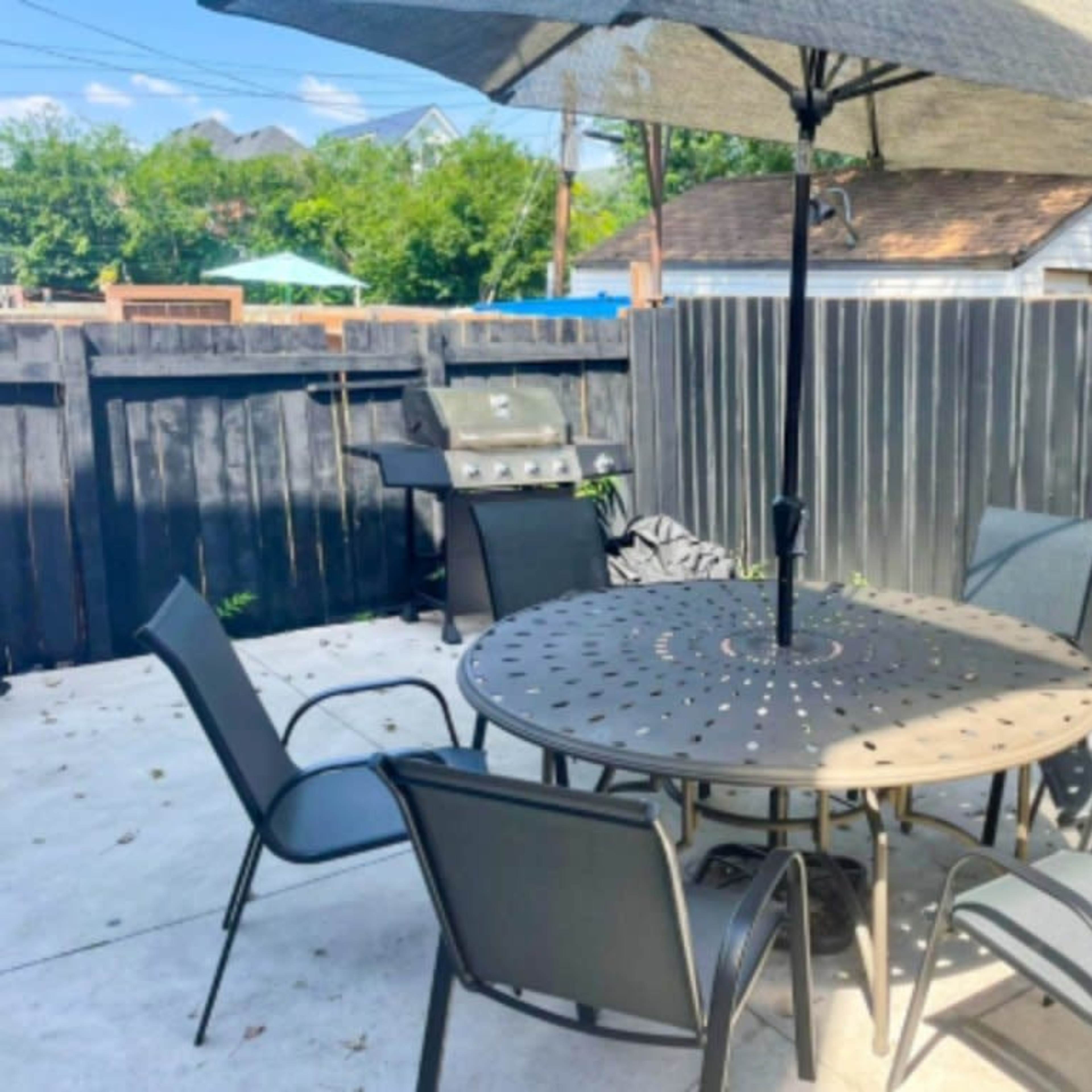 A patio area features a round table with six chairs under a large umbrella, adjacent to a grill and surrounded by a wooden fence.
