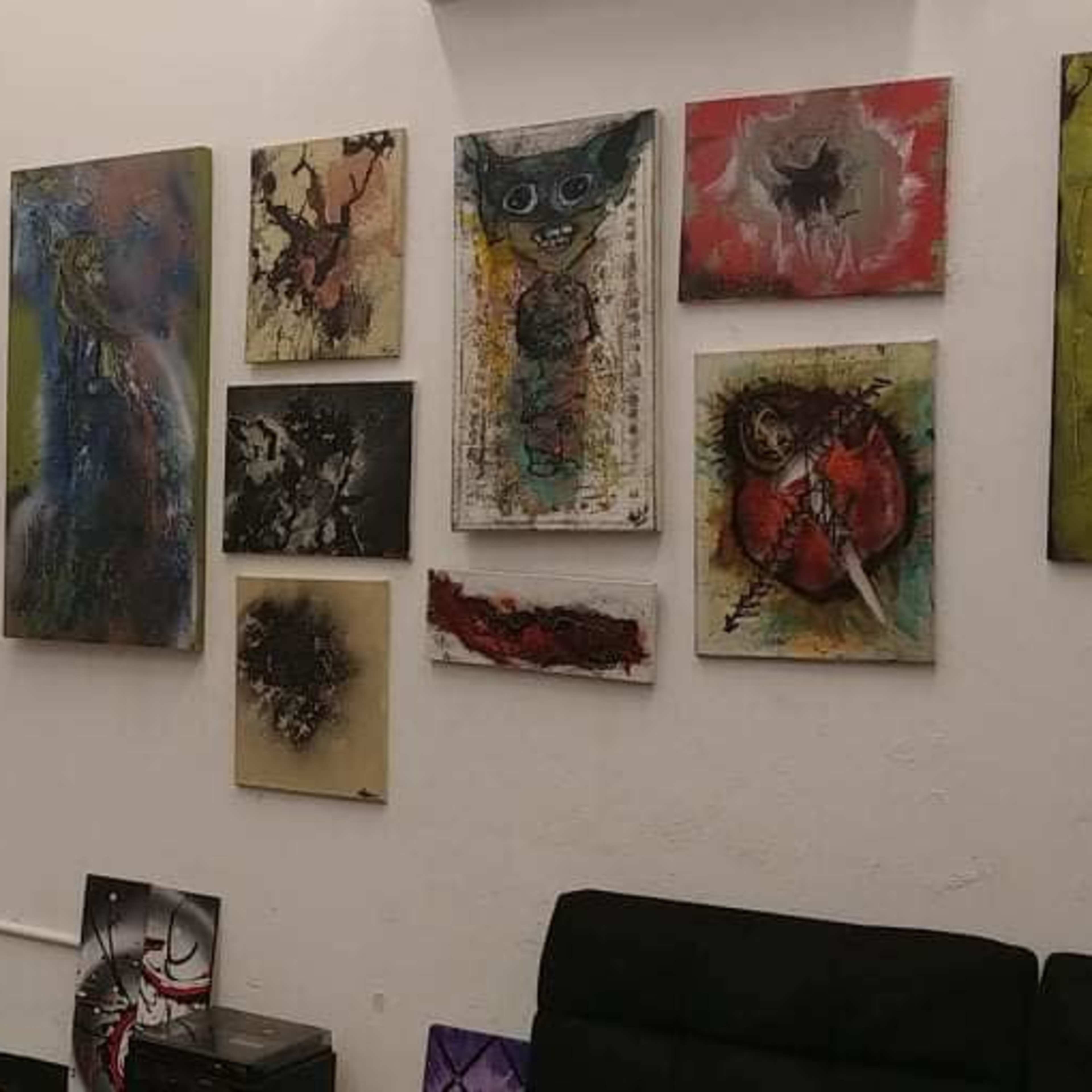 A gallery wall filled with various framed artworks, including abstract and figurative pieces, alongside a dark couch in a simple interior space.