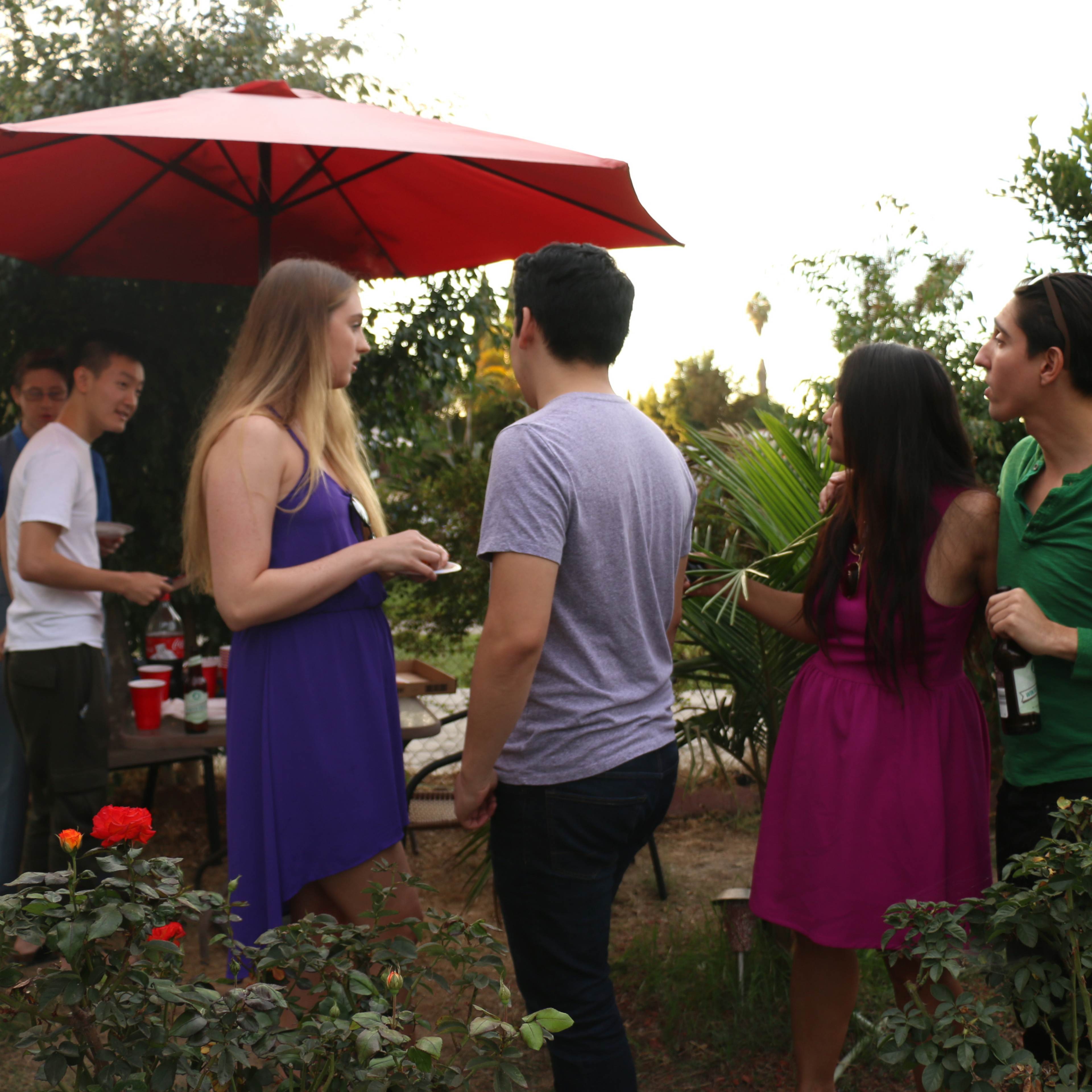 A group of individuals interacts around a patio table under a red umbrella in a backyard garden.
