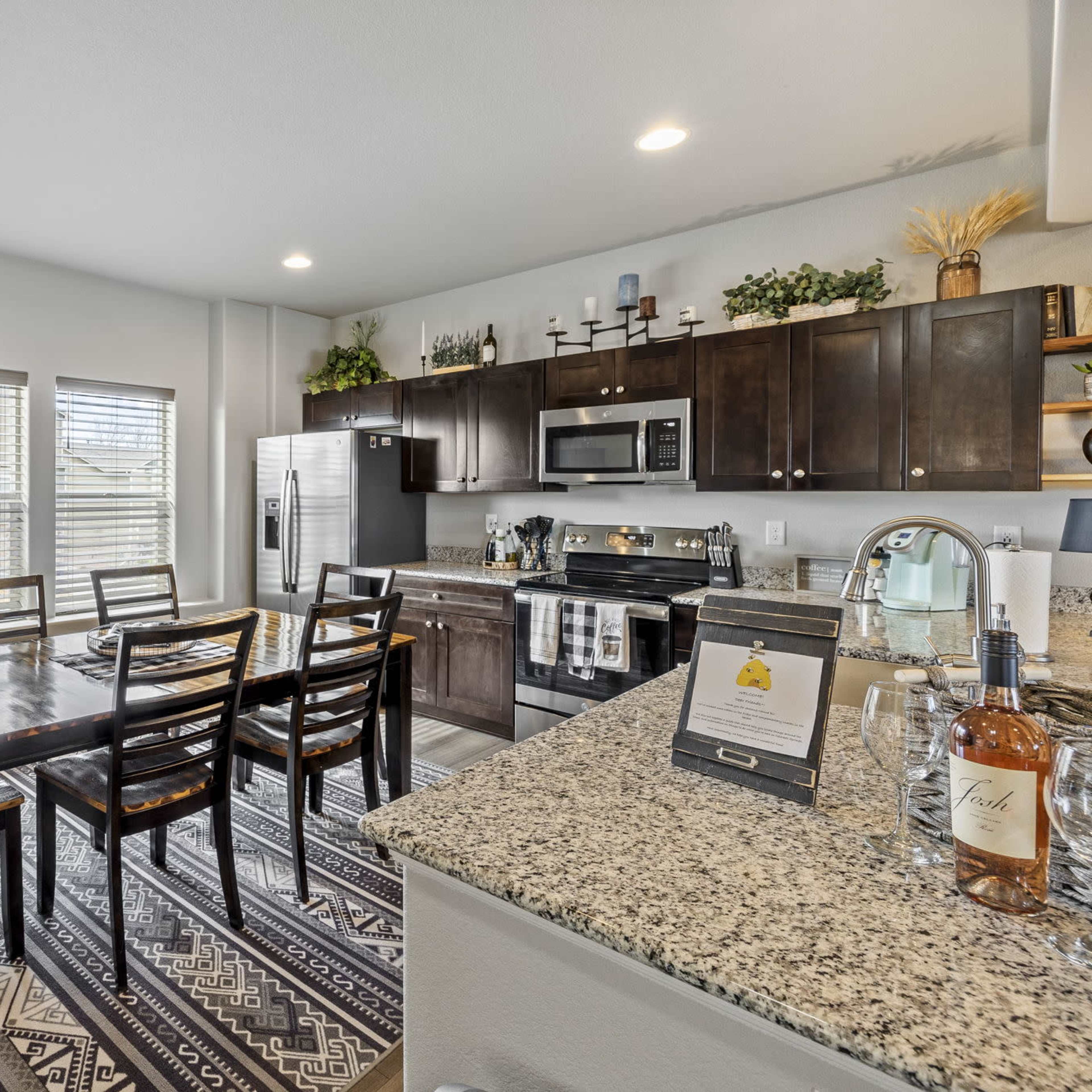 A modern kitchen with dark wood cabinetry, a large dining table, and stainless steel appliances is arranged in a well-lit space with a patterned rug.
