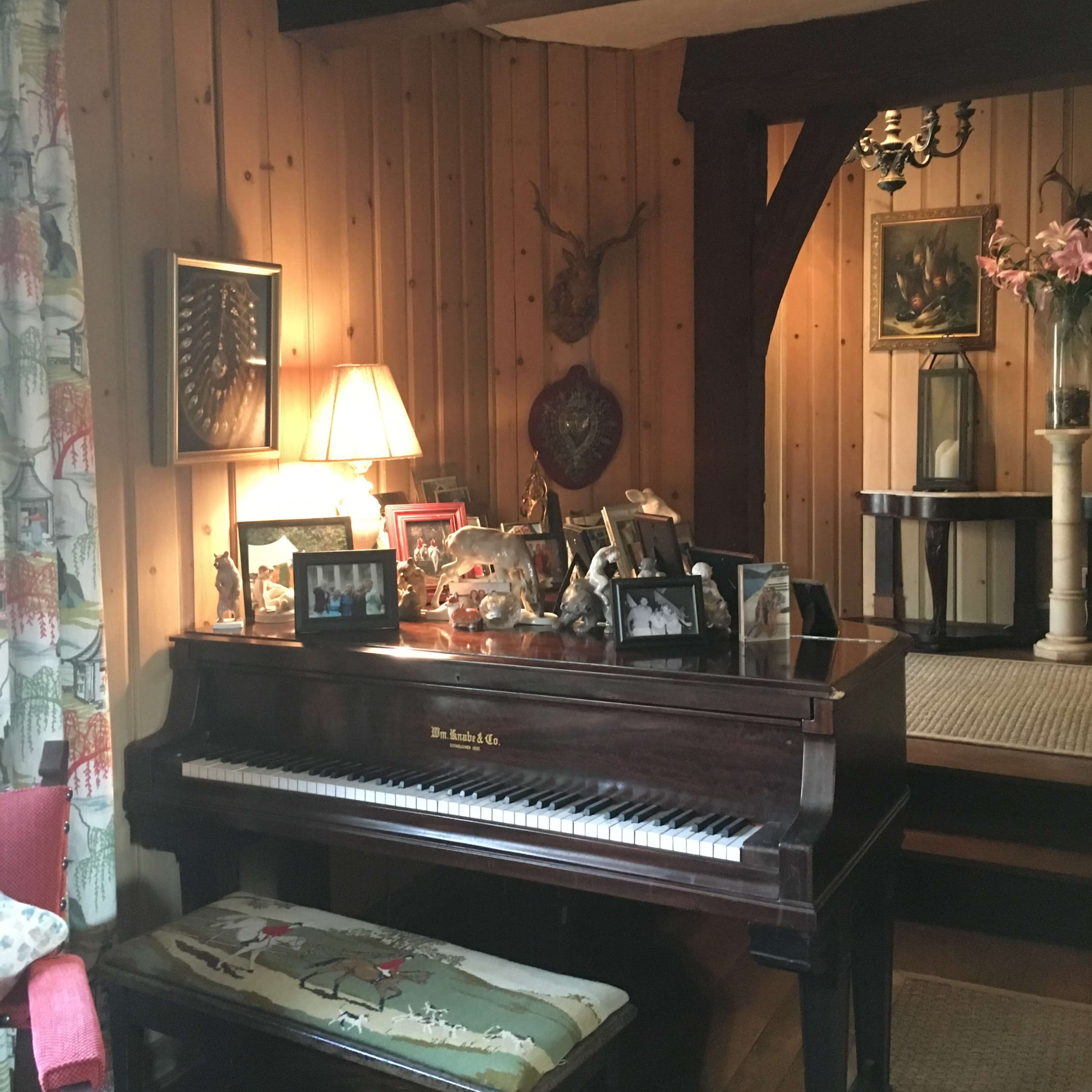 An upright piano with framed photos and decorative items sits against a wooden wall in a cozy room.