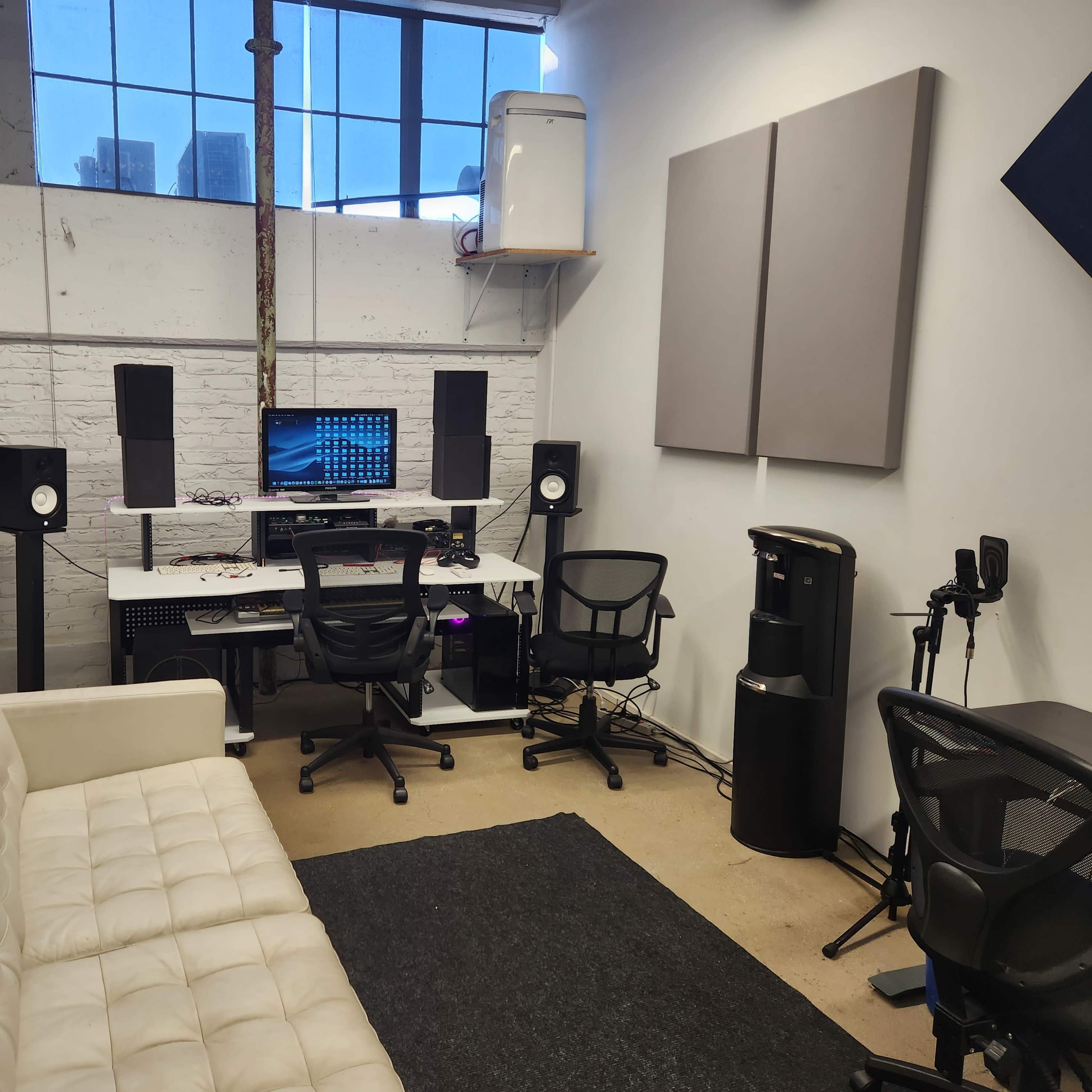 A small audio recording studio with a couch, two office chairs, a computer setup, and soundproofing panels on the walls.