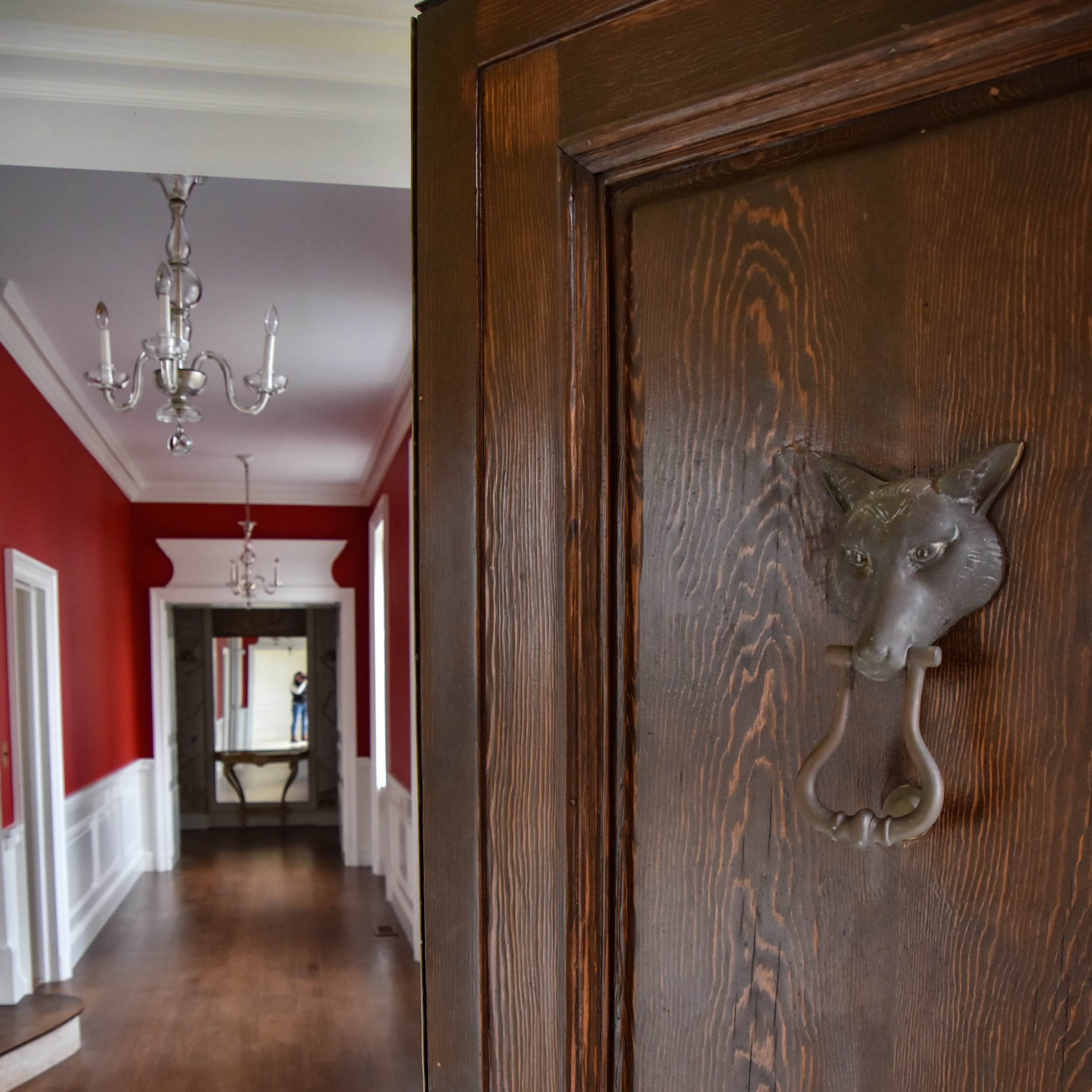 A wooden door with a fox-shaped knocker opens to a hallway featuring red walls and a mirror at the far end.