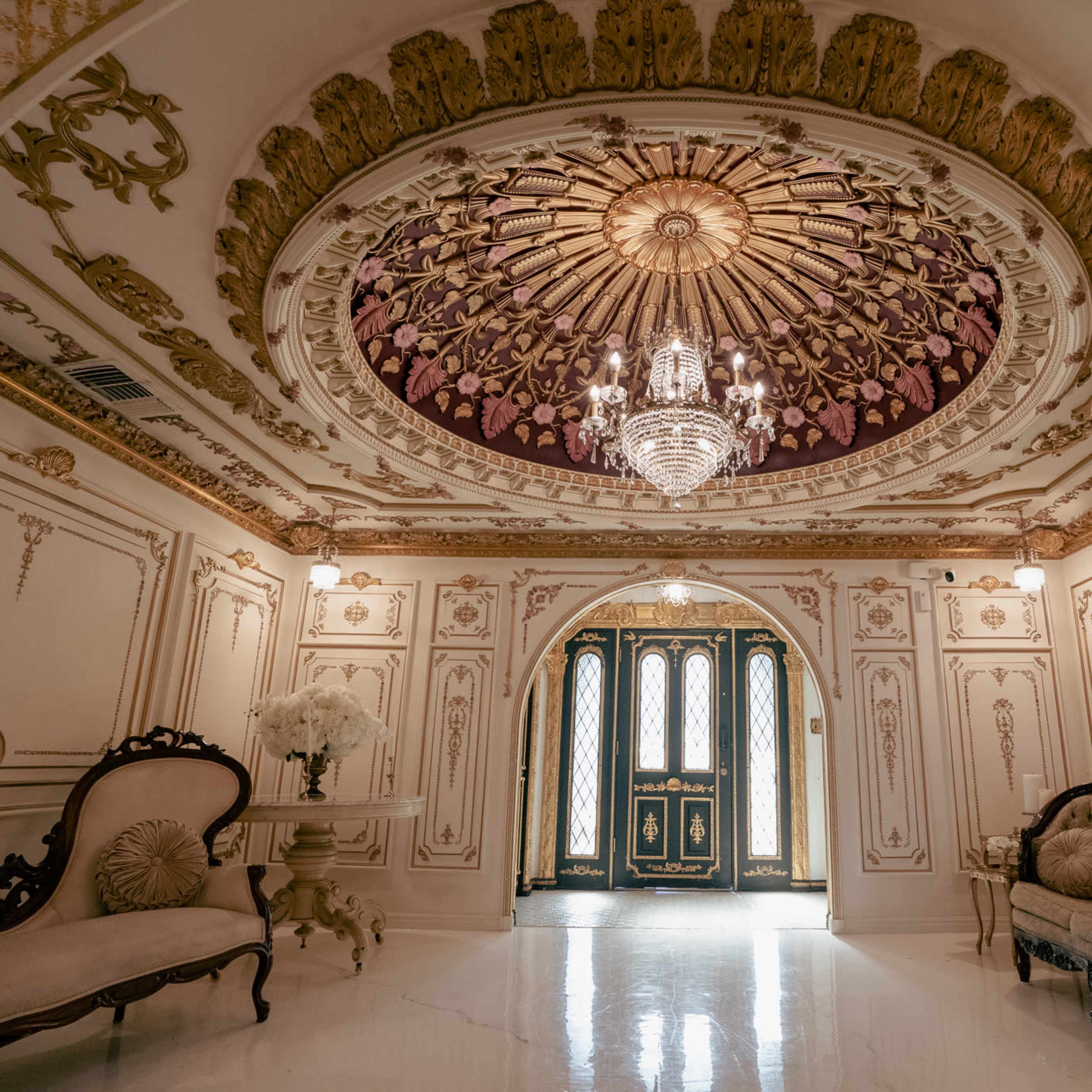 An ornate foyer with a chandelier, detailed ceiling, and ornate furniture, leading to a set of double doors.