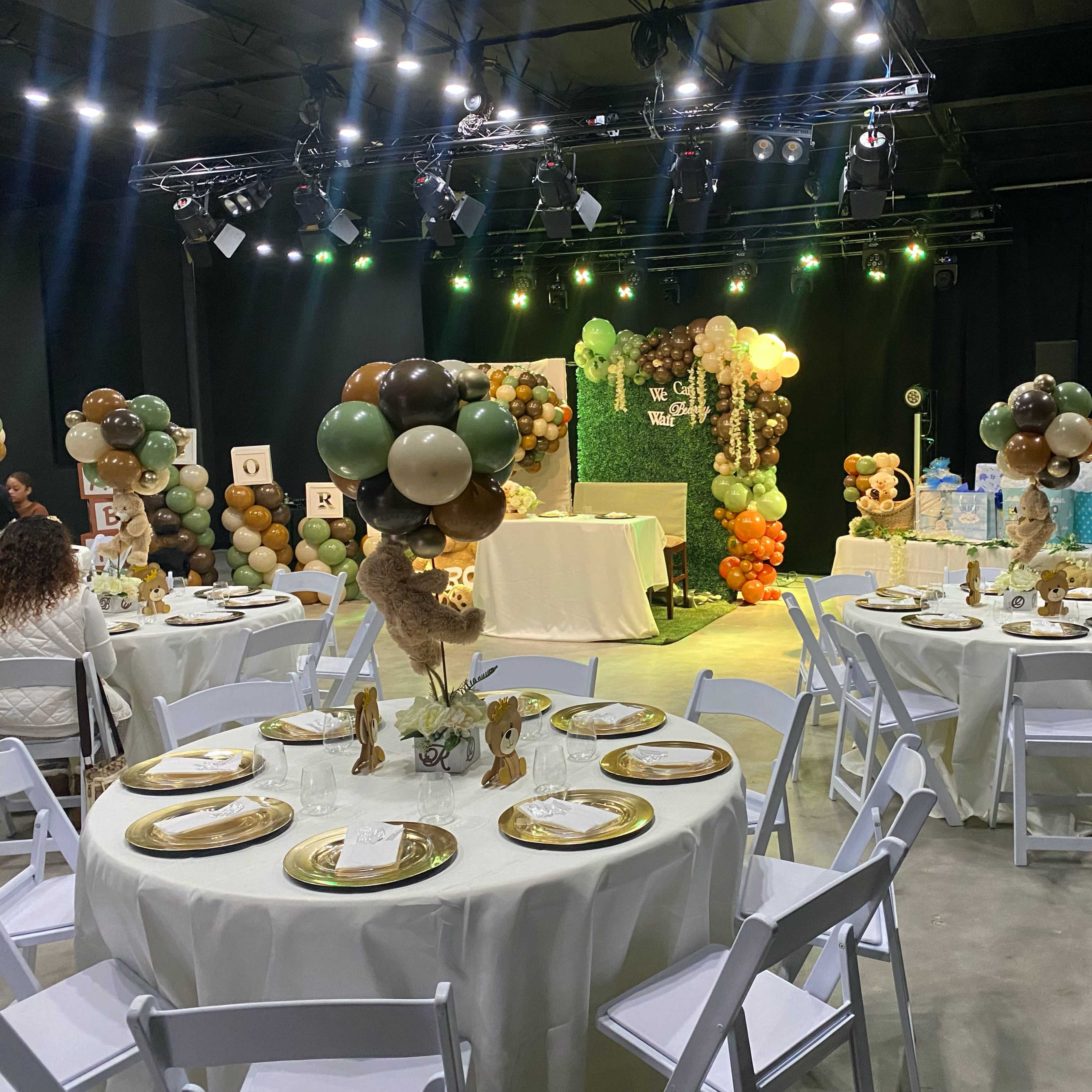 A decorated event space with round tables set for a gathering, featuring balloon displays and a backdrop adorned with greenery and decorative elements.