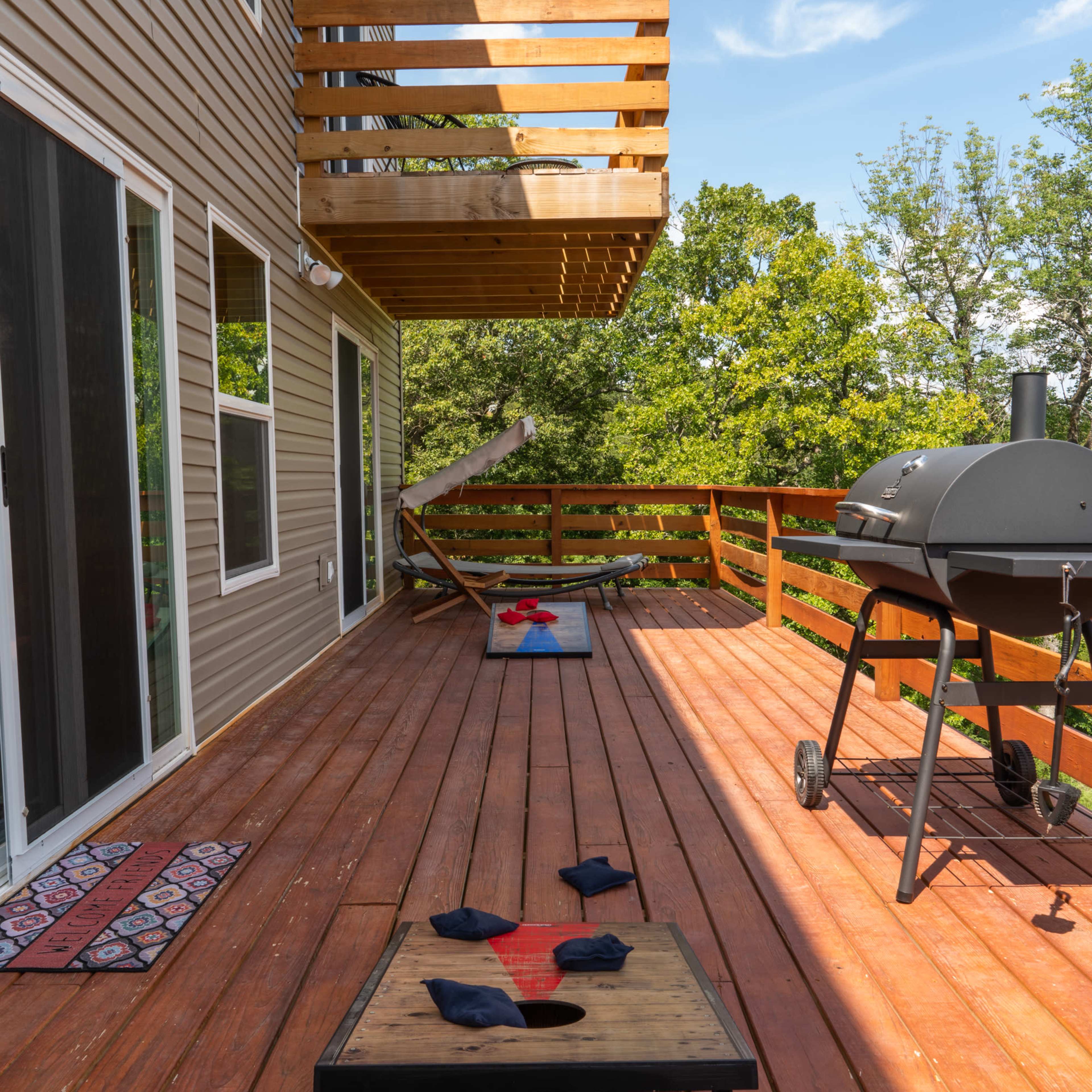 A wooden deck with two bean bag toss boards, a charcoal grill, and a balcony overlooking the yard.