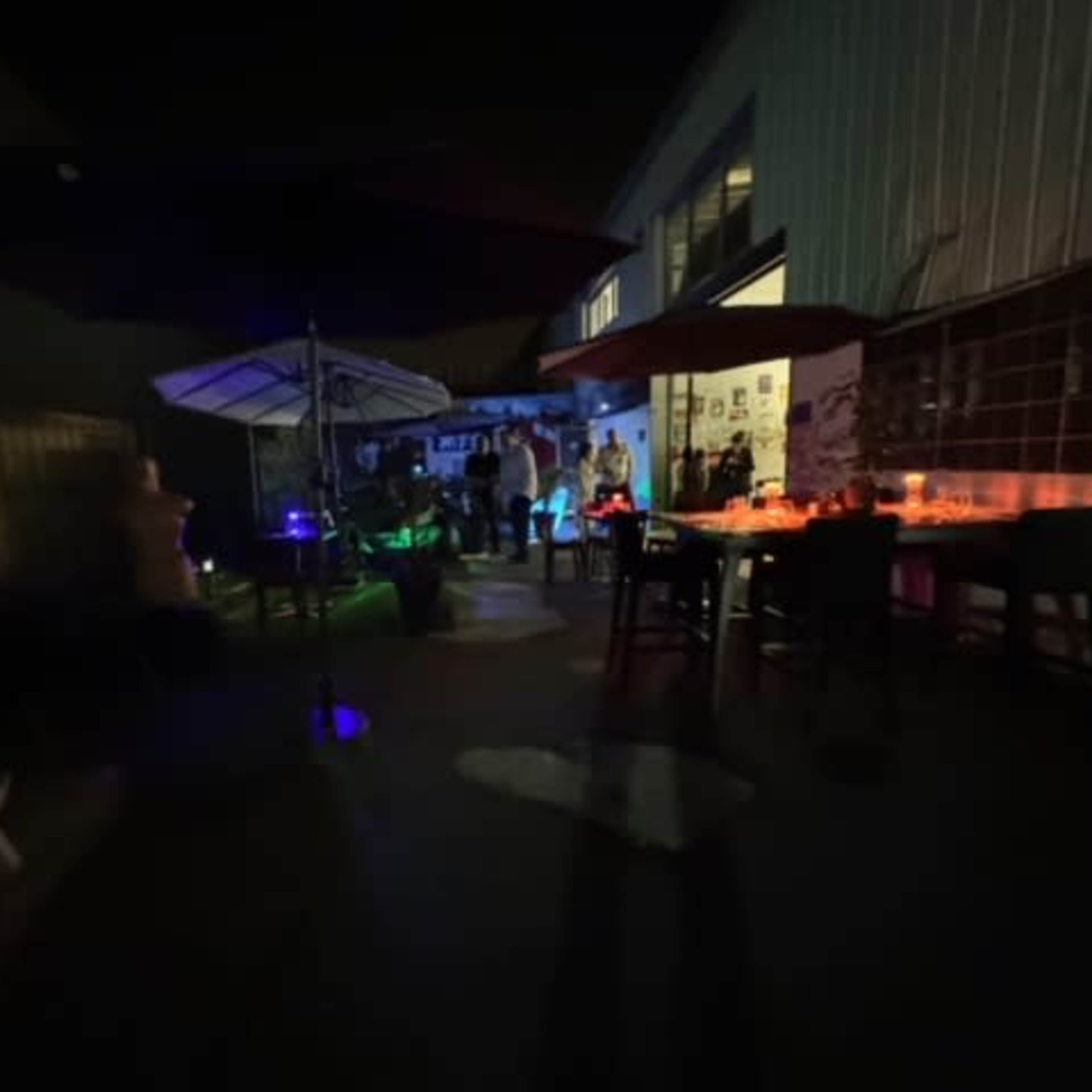 A dimly lit outdoor gathering space with tables, chairs, and colorful lighting, featuring several people in the background.