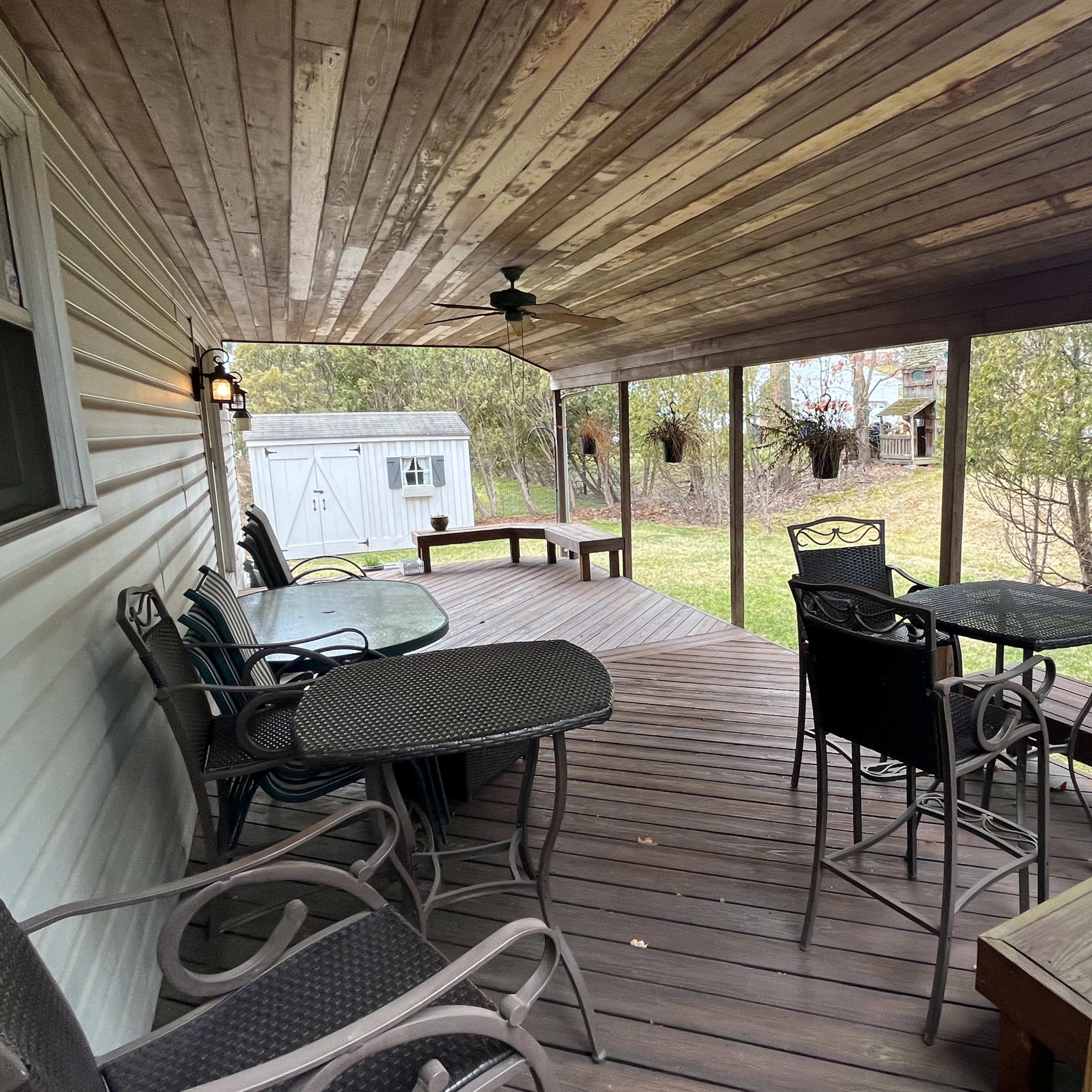 A covered porch with wooden flooring, featuring several outdoor chairs and tables, along with a shed visible in the background.