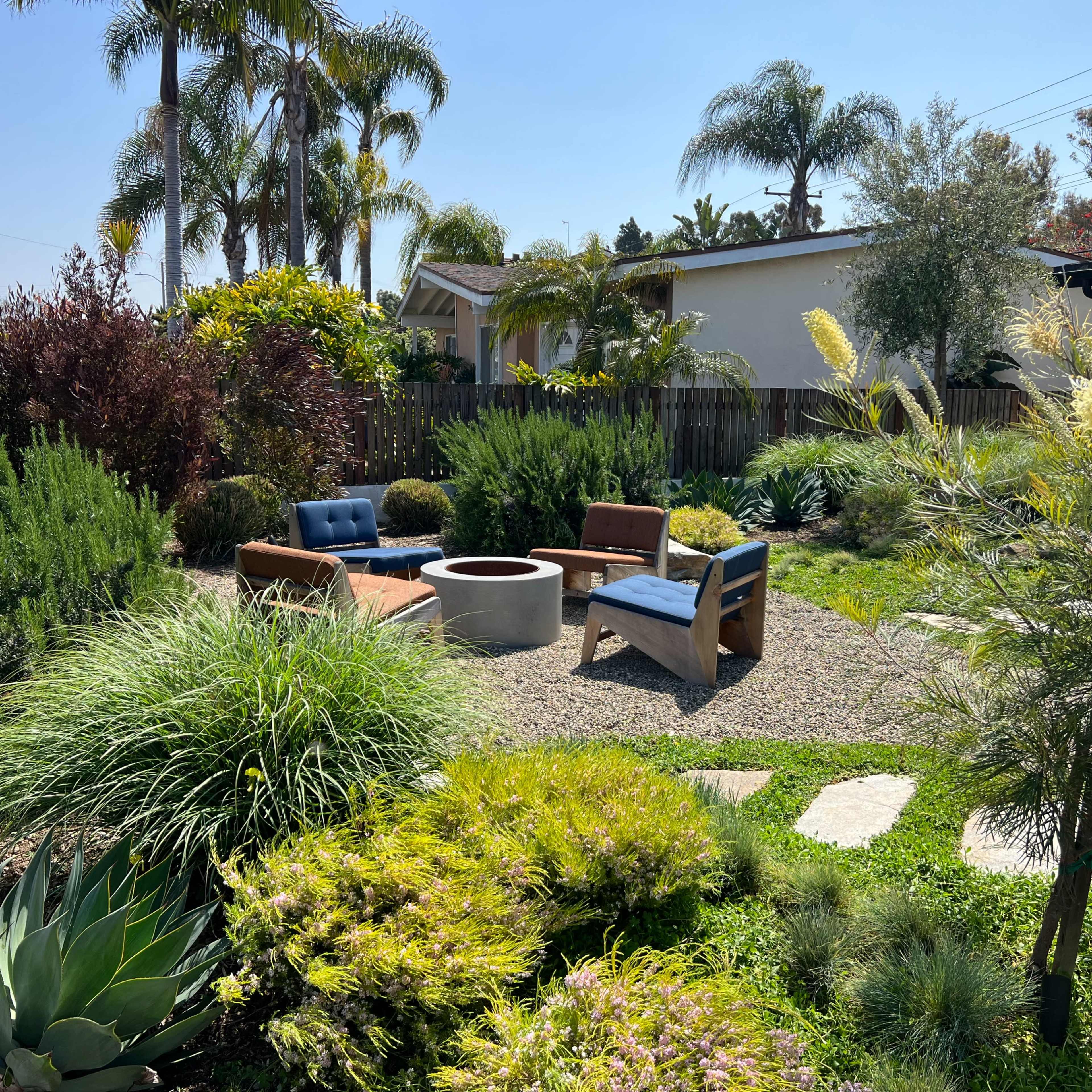 A landscaped garden featuring a circular fire pit surrounded by several chairs and diverse plants, including palms and shrubs.