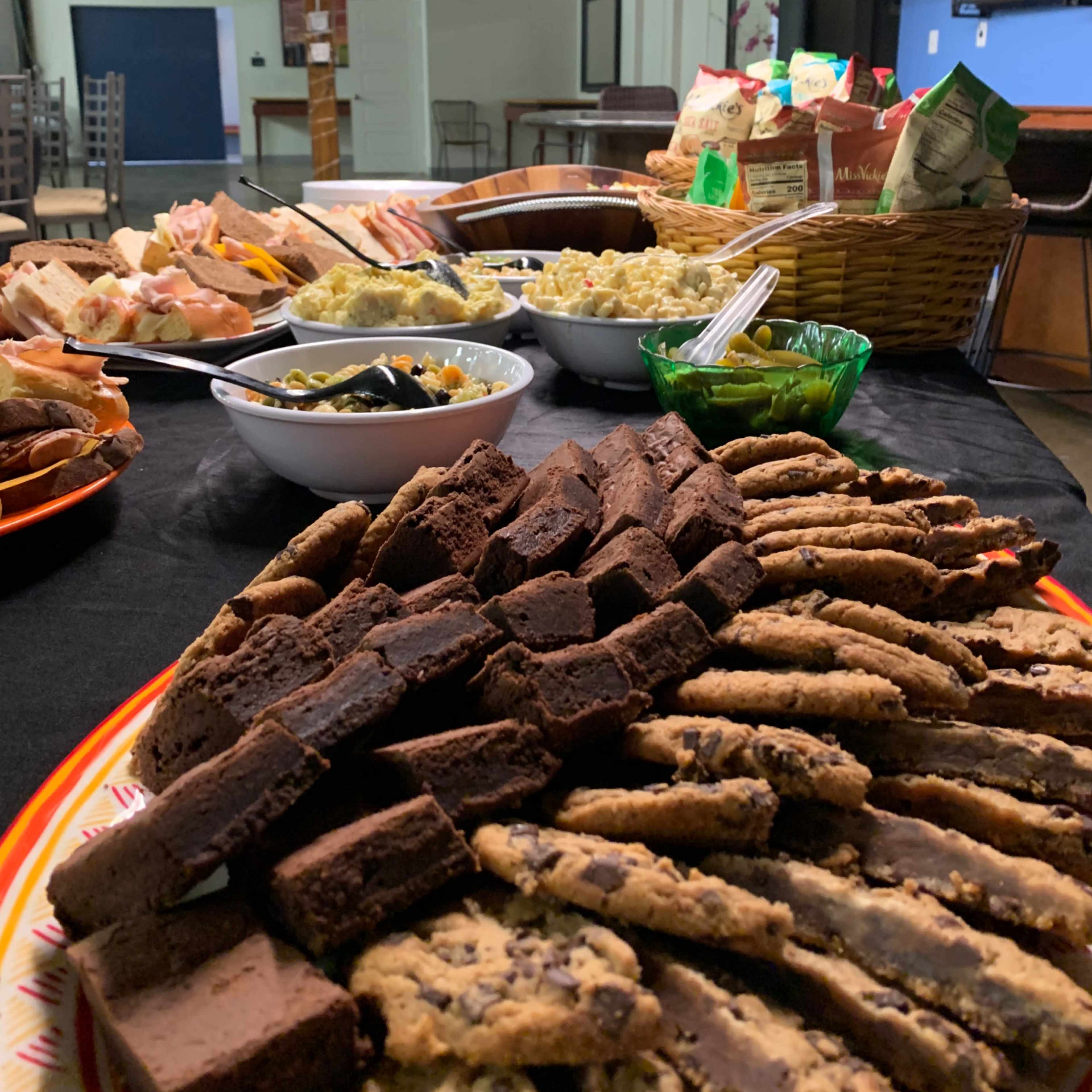 A table is set with various dishes, including cookies, brownies, sandwiches, salads, and snacks in a spacious indoor setting.