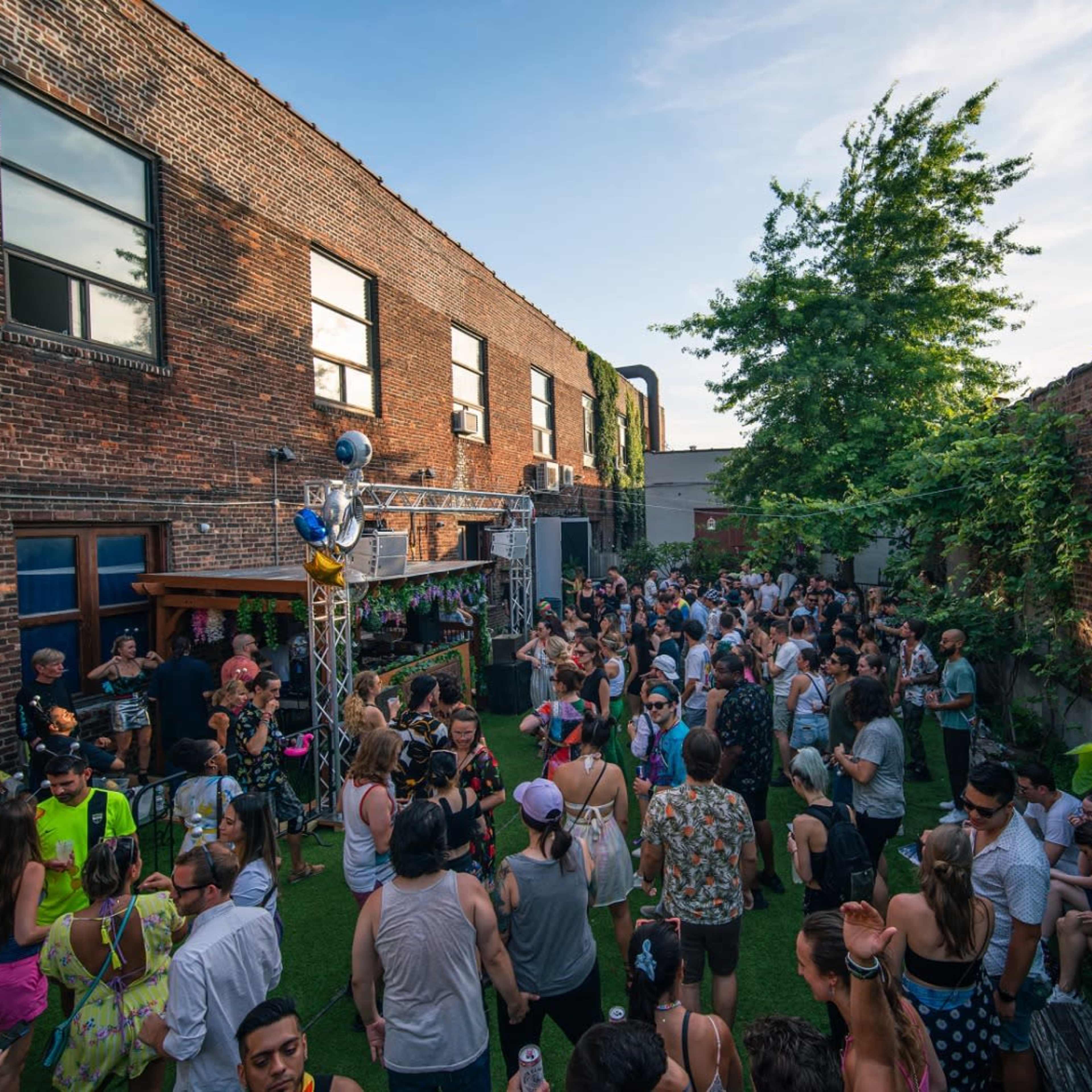 A large crowd of people gathers in a vibrant outdoor space surrounded by brick walls and greenery, enjoying a lively event.