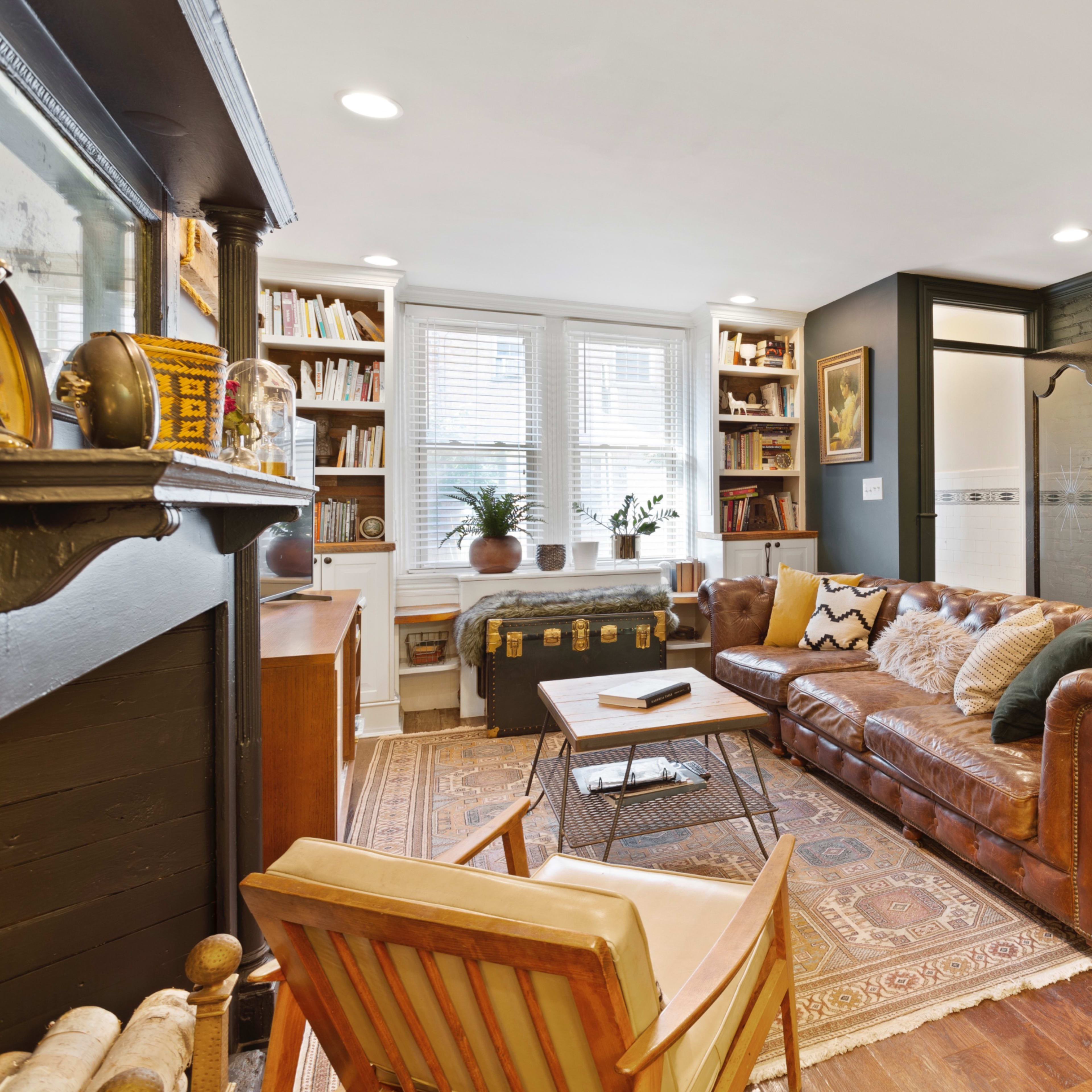 A cozy living room featuring a brown leather sofa, a wooden armchair, a bookshelf, and decorative items against a dark green wall.
