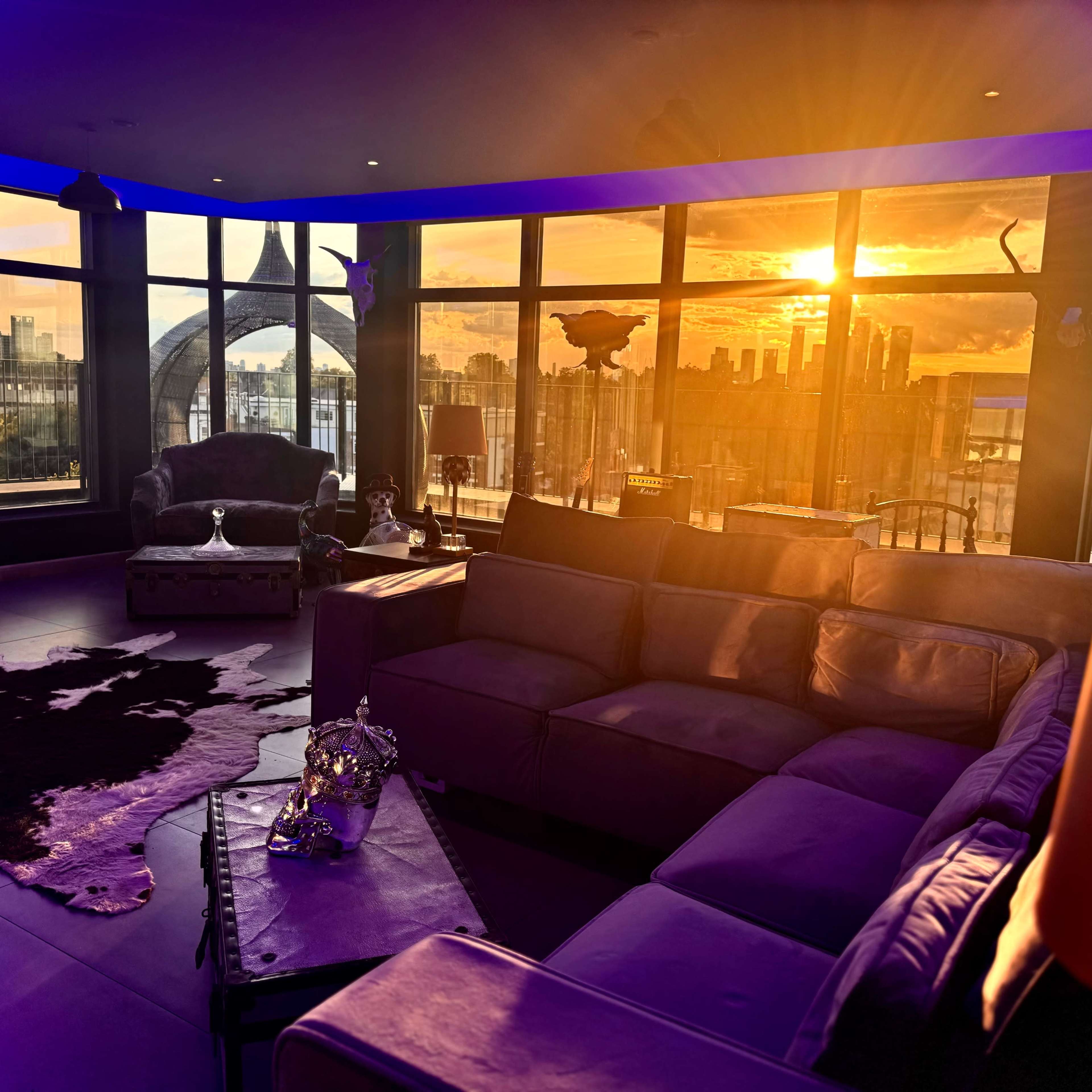 A modern living room features a sectional sofa, large windows displaying a sunset over a city skyline, and decorative accents including a cowhide rug.