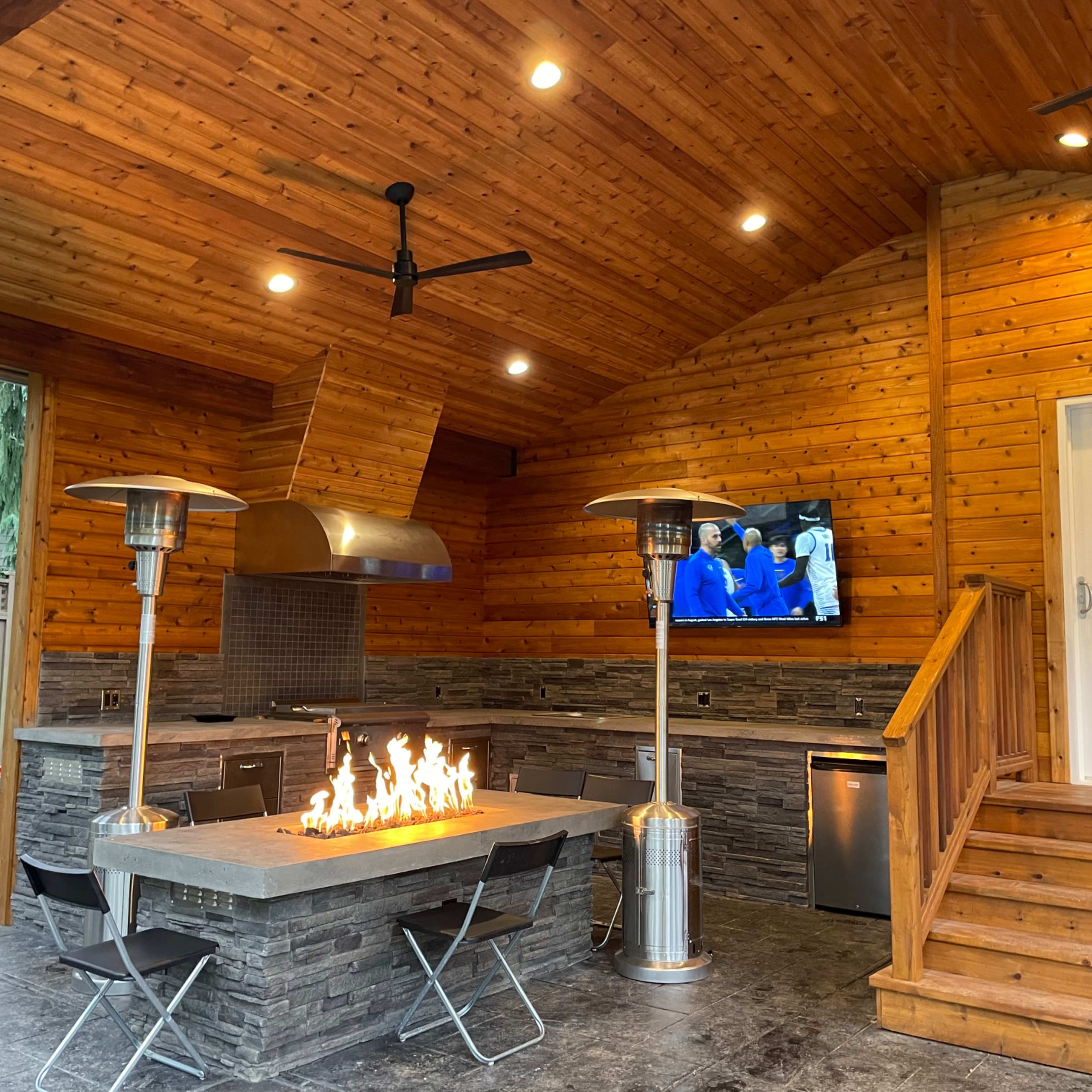 A modern outdoor kitchen with a stone countertop, a built-in grill, two patio heaters, and a television mounted on the wooden wall.