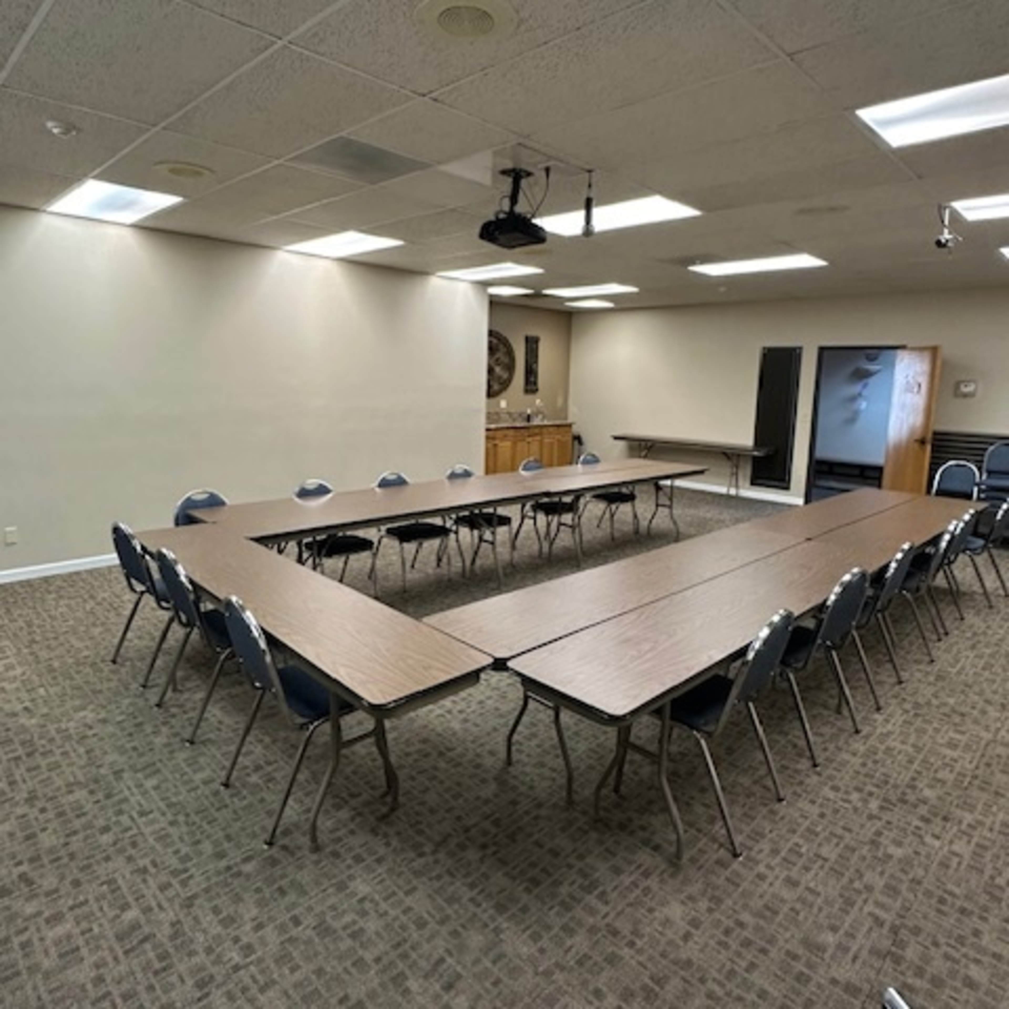 A conference room with two long tables arranged in a U-shape and chairs placed around them, featuring a projector overhead.
